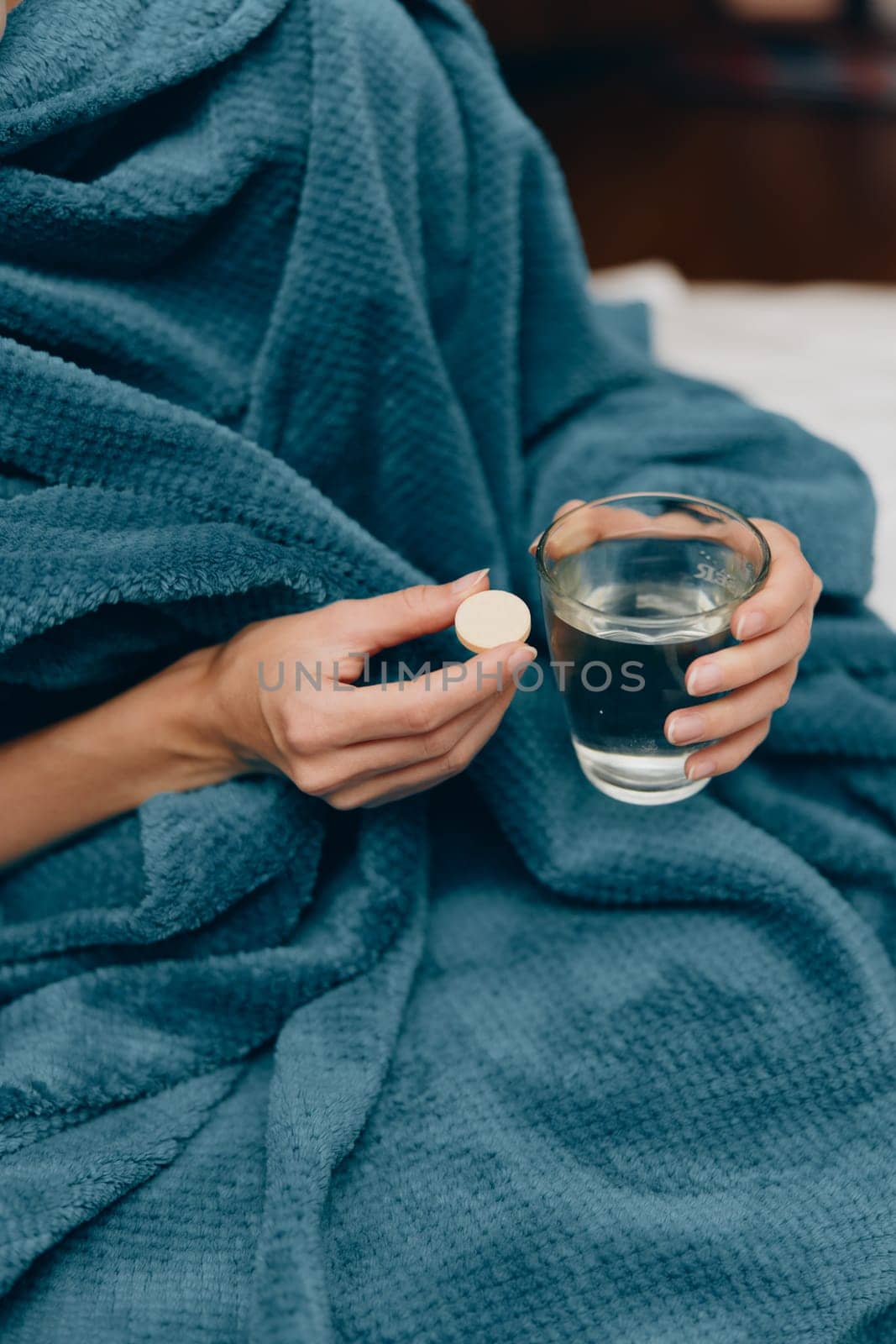 Woman in blue blanket sitting on bed, holding glass of water and pill in hand.