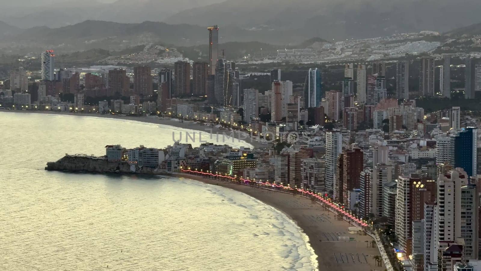 A picturesque view of a modern coastal city with illuminated skyline and tranquil waters during sunset.