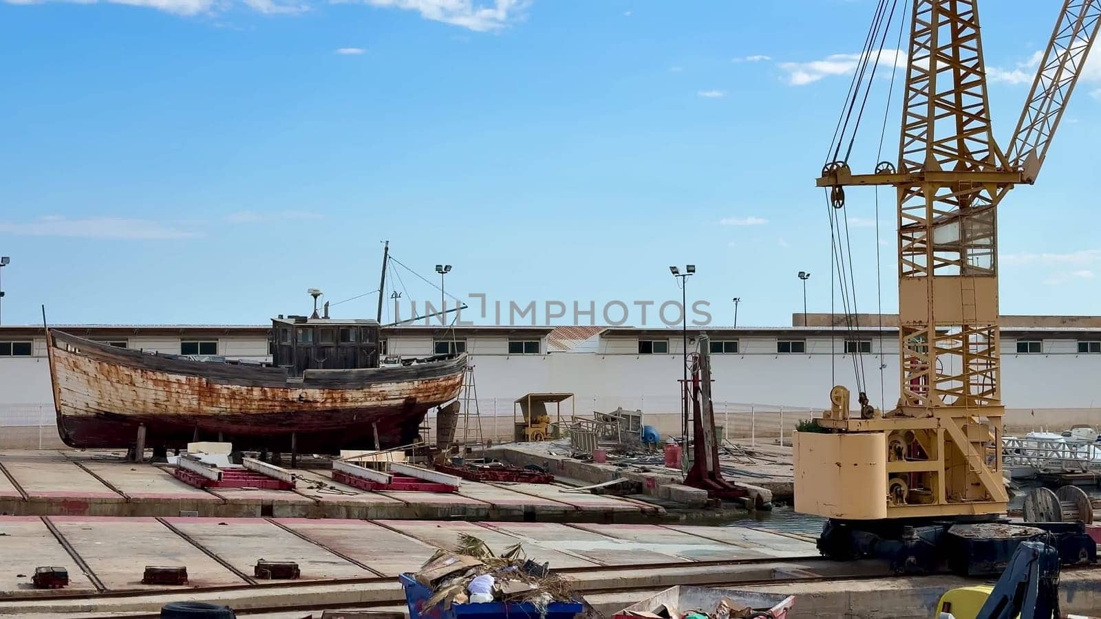 An old and weathered fishing boat now sits in a shipyard, accompanied by a yellow crane ready for its restoration