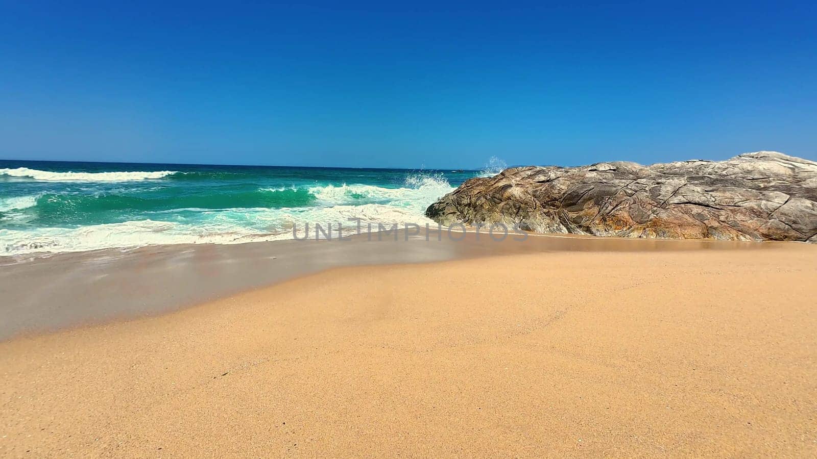 A picturesque beach scene featuring soft sand, gentle waves, and a large rock formation under a clear sky.