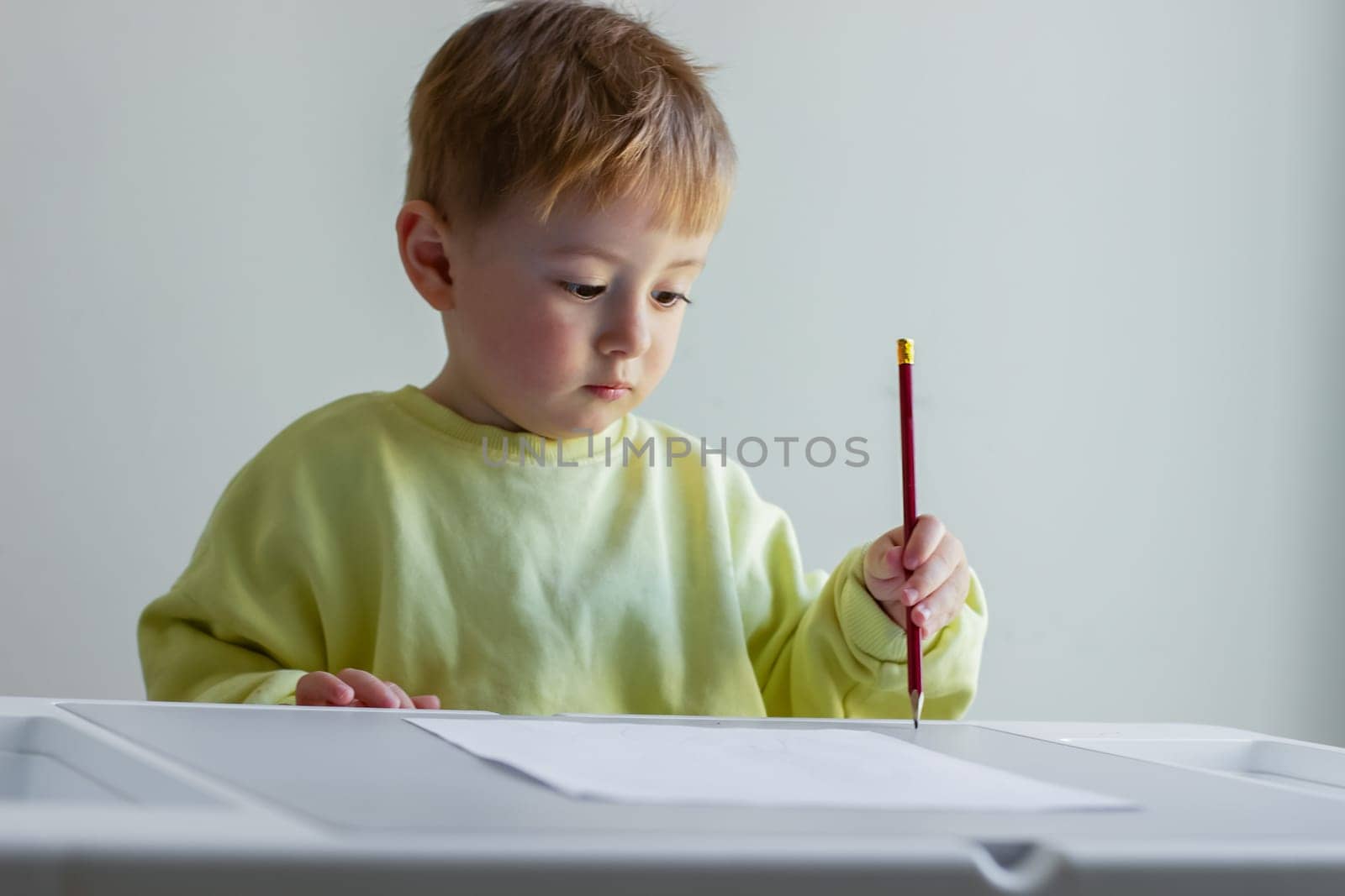 Cheerful little boy in a yellow sweater draws with colored pencils on paper at a table. High quality photo