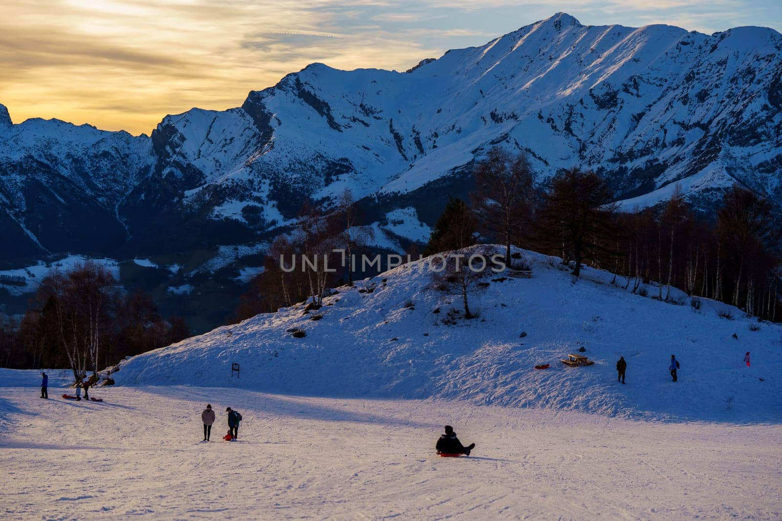 Piani di Bobbio, Italy - 24.12.2022: People enjoy sledding on snow-covered hill in Bobbio with stunning backdrop of snow-capped mountains illuminated by golden sunset