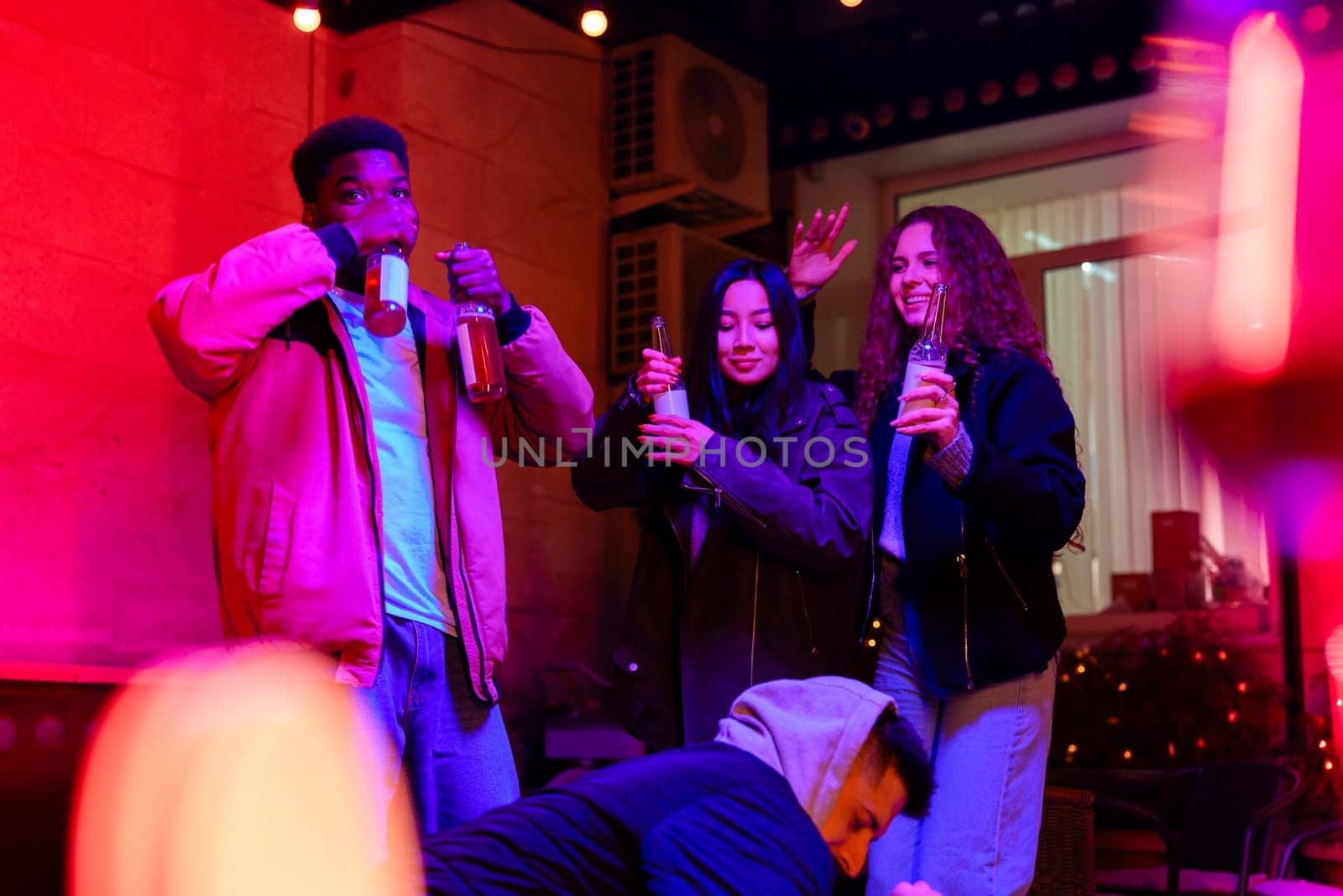 A group of friends celebrates together, clinking their drinks and dancing joyfully in a warm, inviting indoor space illuminated by colorful lights. The atmosphere radiates fun and camaraderie.