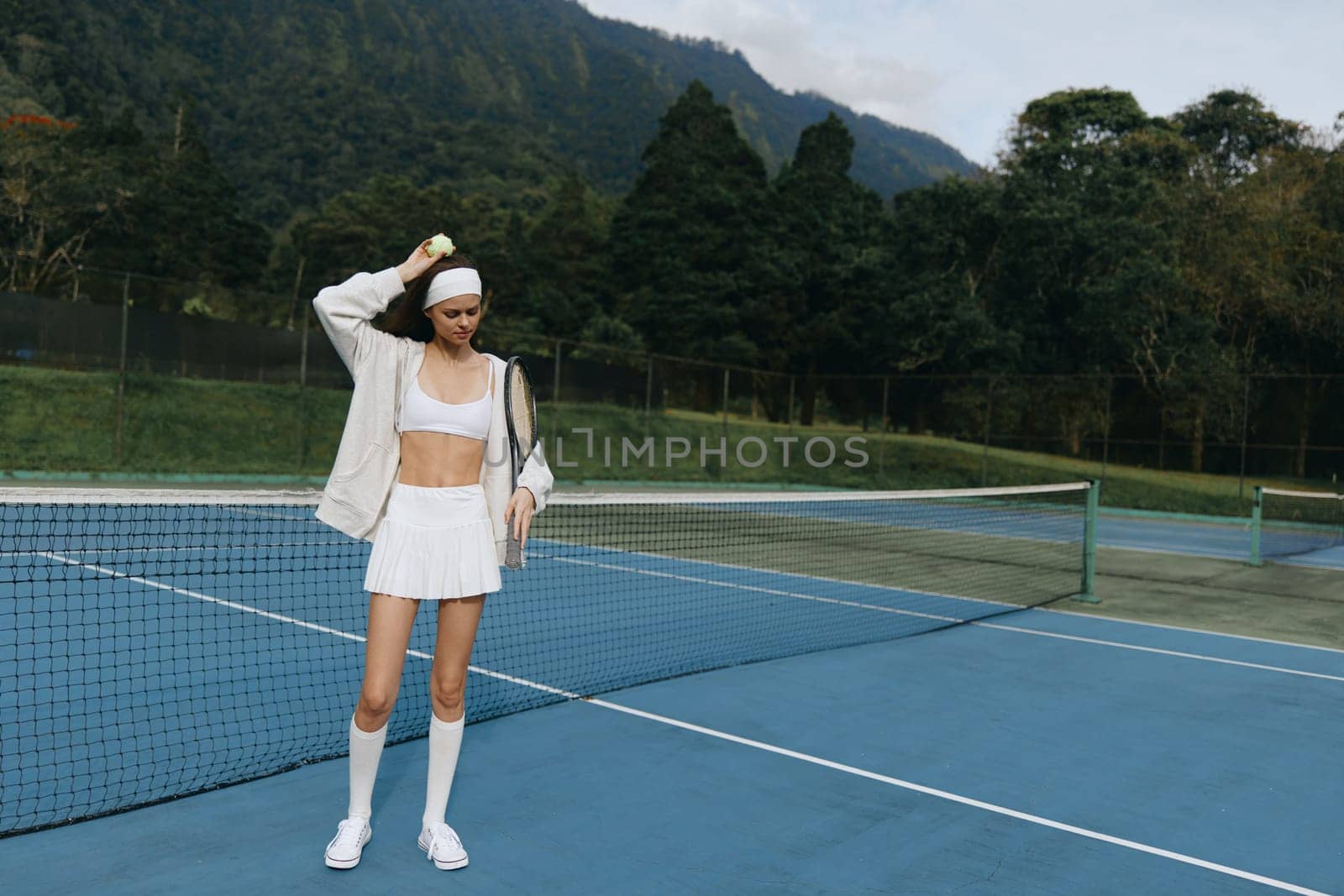 Athletic woman in a white sports outfit playing tennis outdoors, exuding confidence and energy against a green mountain background, ideal for active lifestyle themes