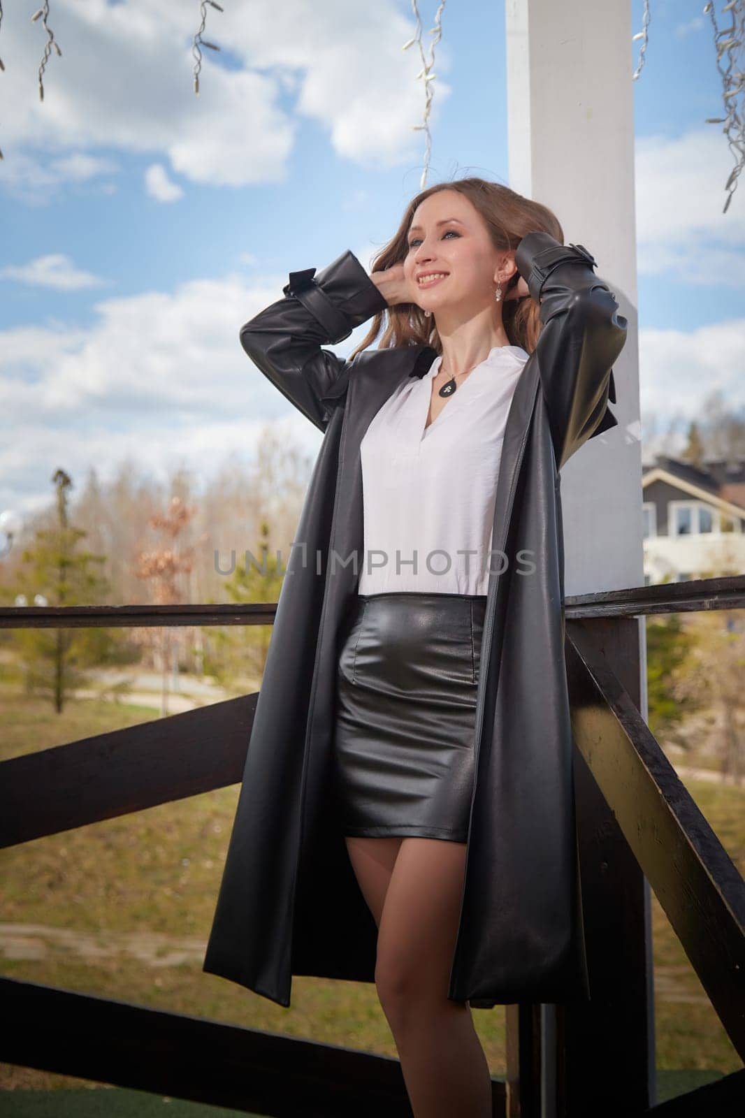A woman in a black leather outfit poses outdoors on a sunny day with trees and a house behind her.