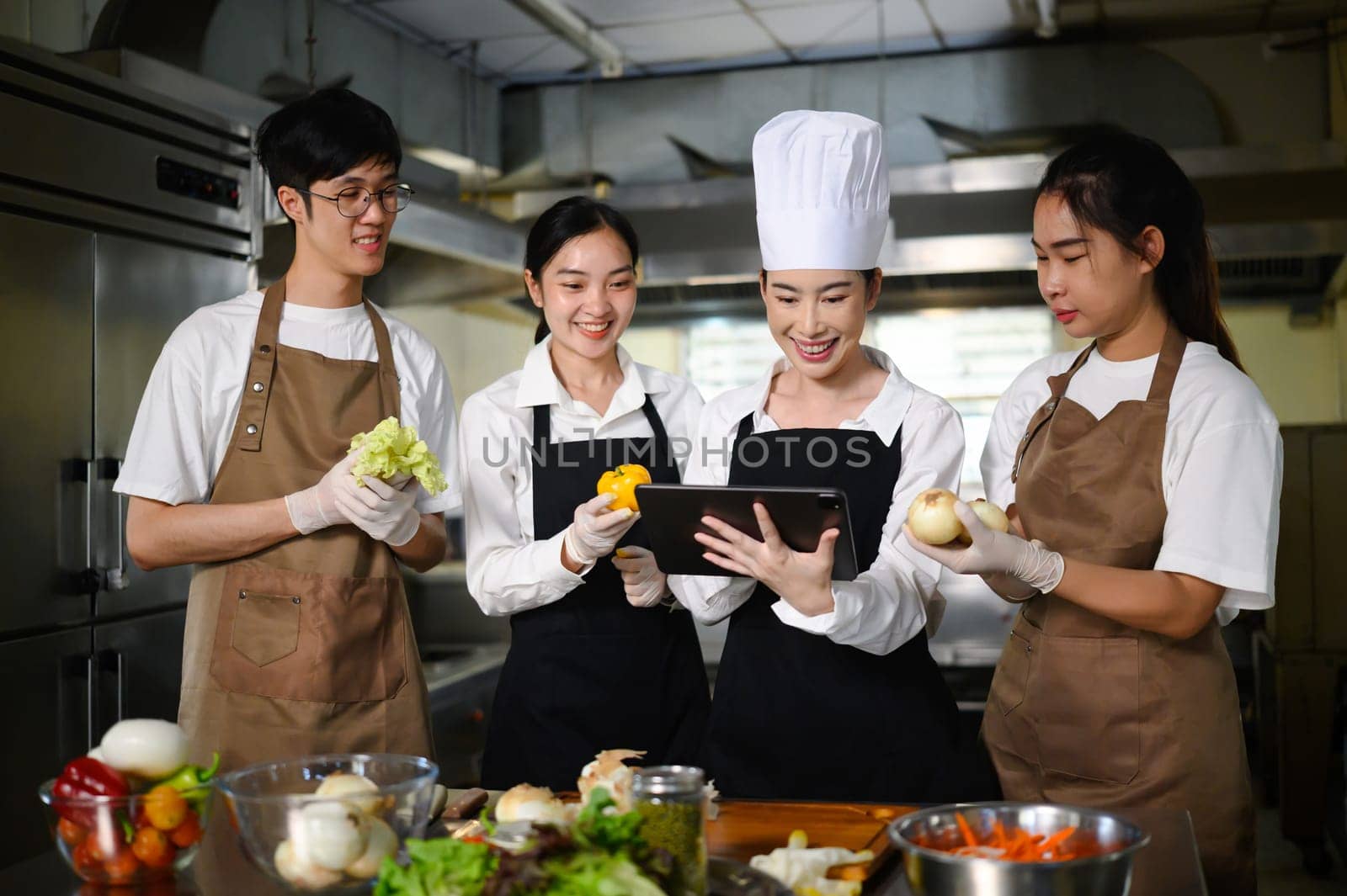 Smiling chef teacher using a tablet to explain a recipe to students in a professional kitchen.