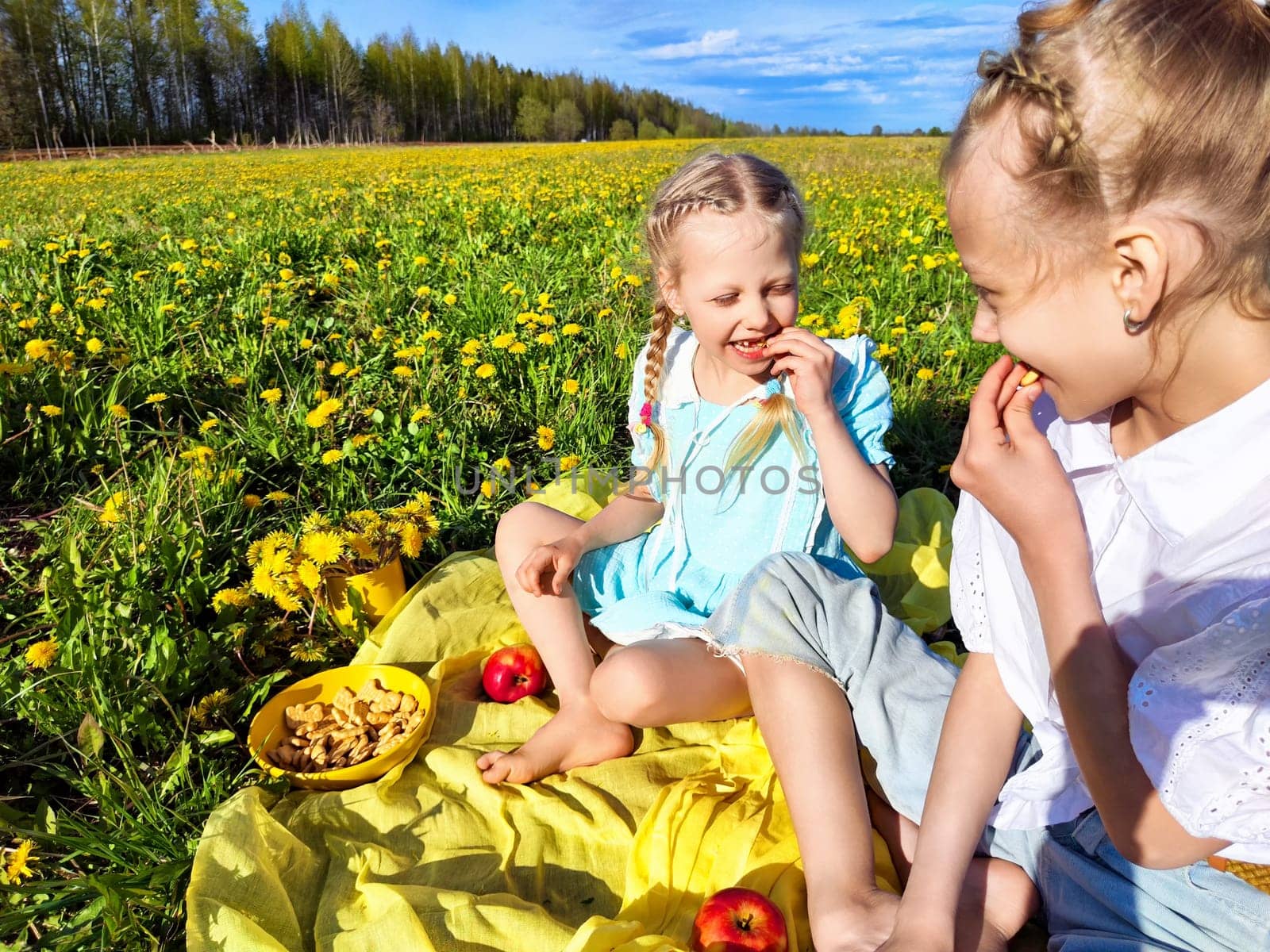 Two young girls share laughter and snacks on a blanket in a colorful dandelion field under a clear blue sky.