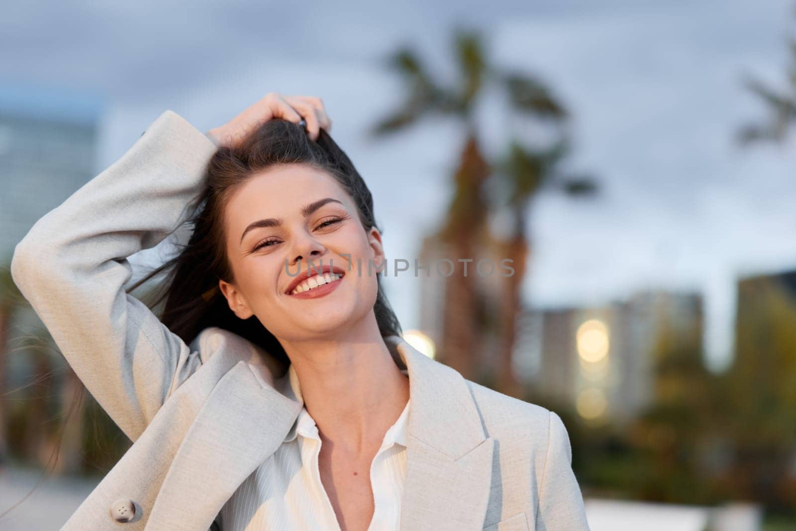 Confident young woman in beige business jacket smiling outdoors with urban cityscape and palm trees blurred in background, natural light enhancing fresh and professional business vibe