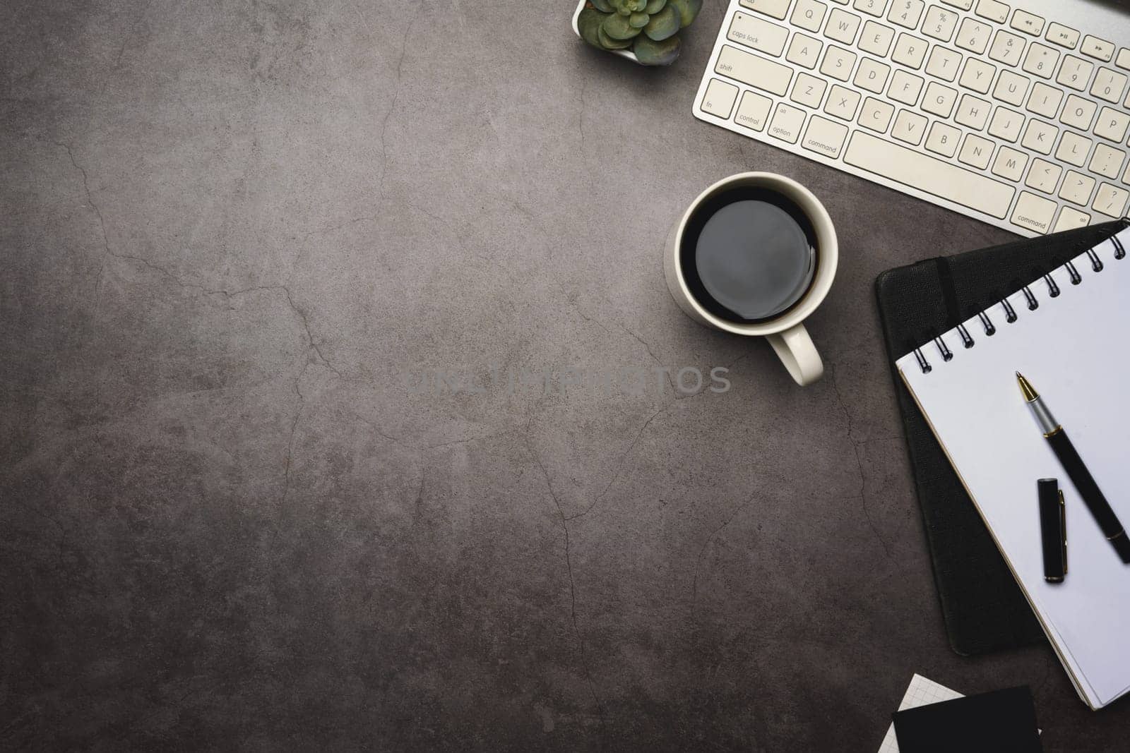 Flat lay of office desk with keyboard, coffee cup and notebook on dark stone surface.