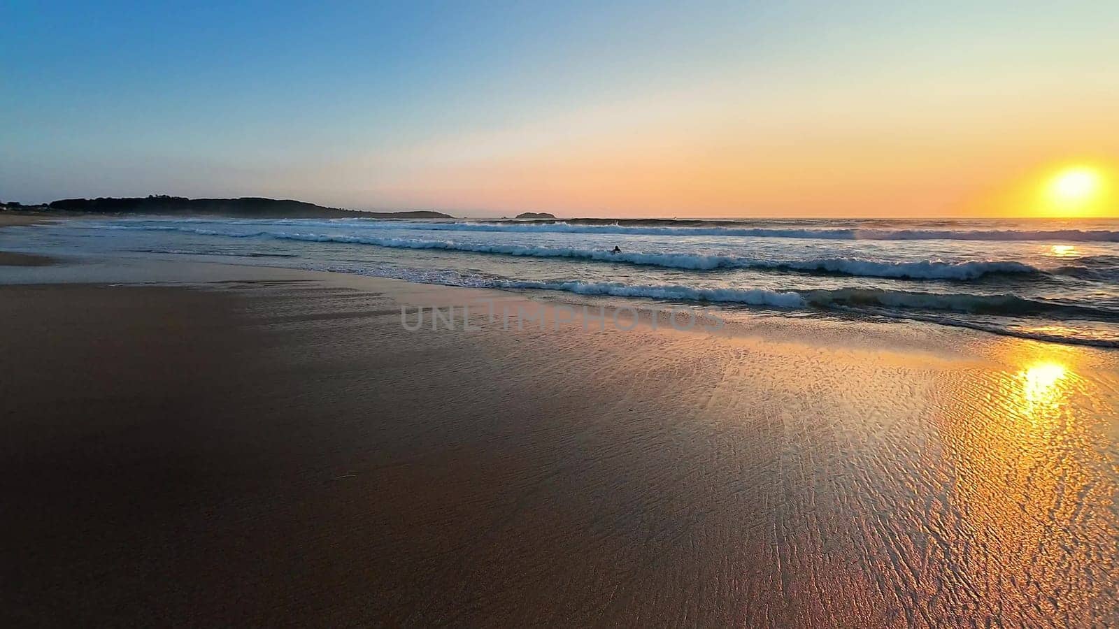 A tranquil beach sunset shows golden reflections on calm waters and soft waves lapping at the shore