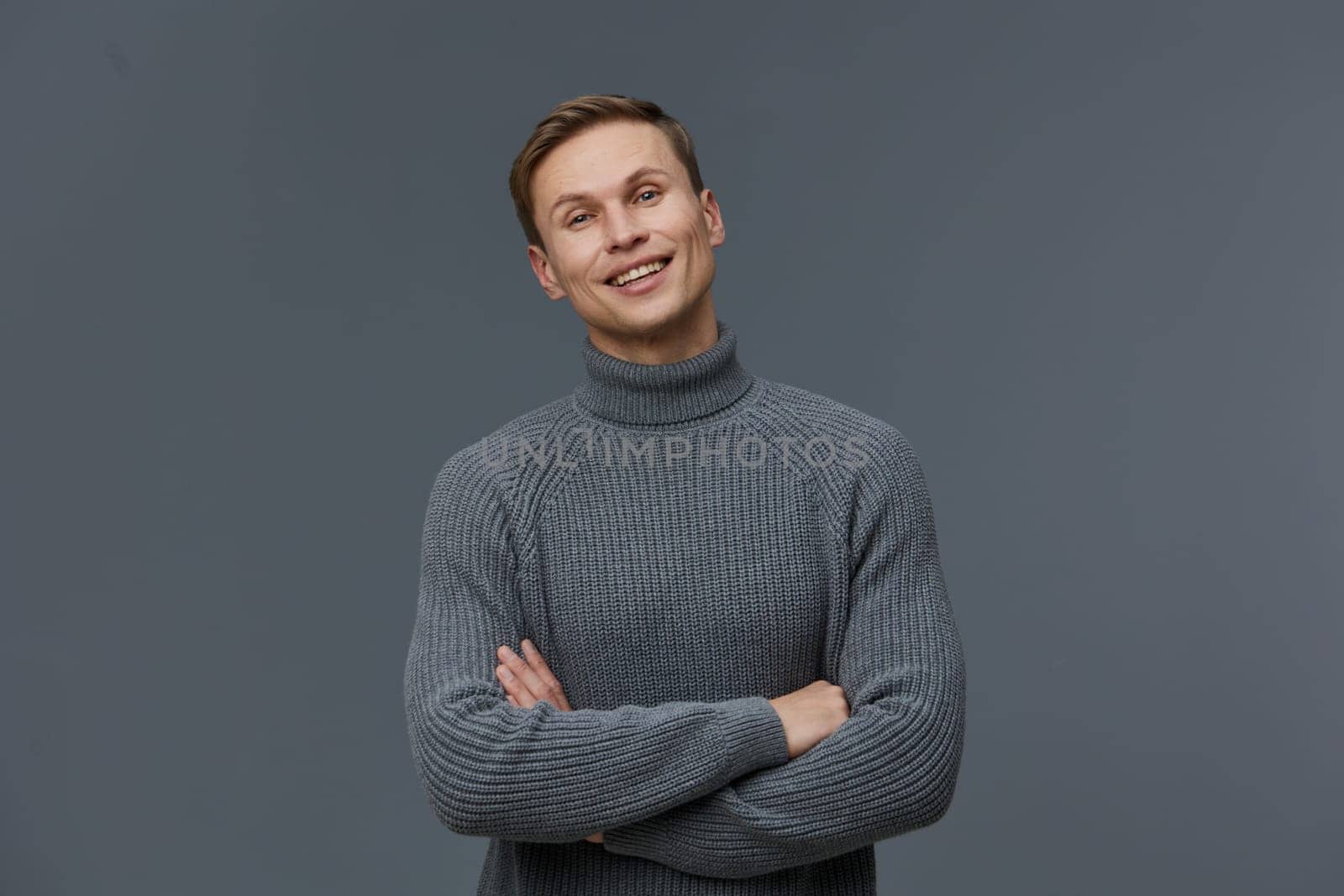 Happy young man wearing a cozy gray turtleneck sweater posing with arms crossed and smiling warmly at the camera, standing in front of a neutral gray background. Concept: confident, casual style, fashion, portrait photography, male.