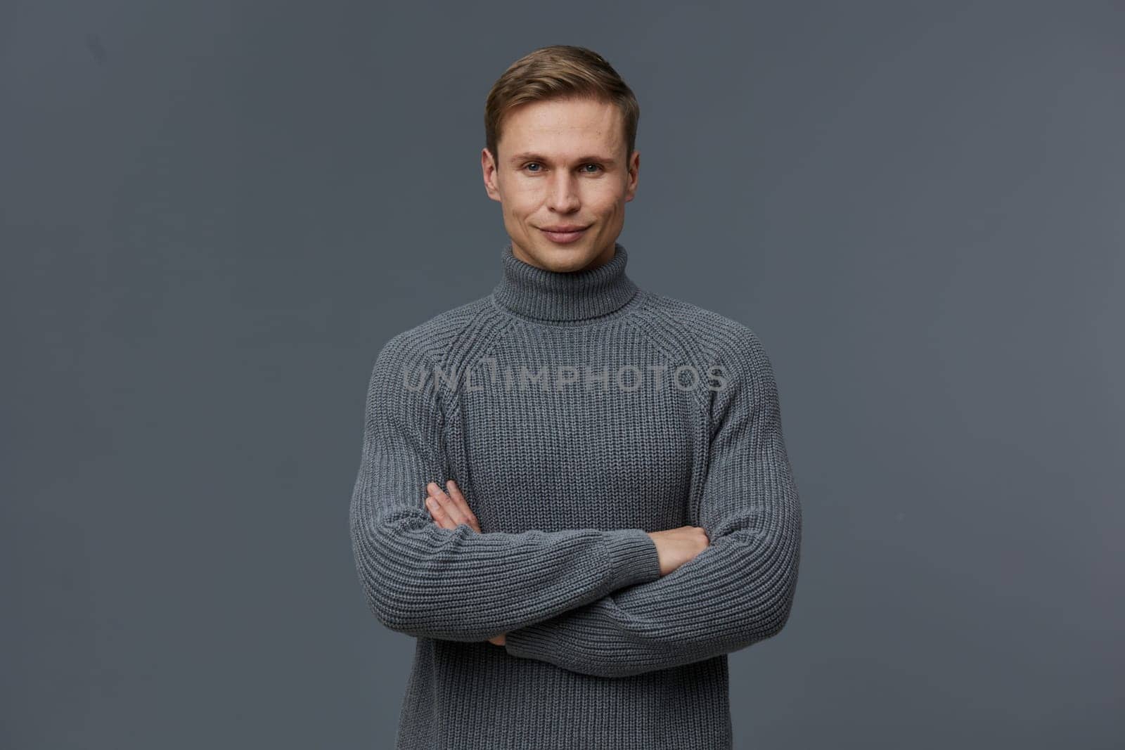 smiling man wearing gray knitted turtleneck sweater standing with arms crossed and looking at camera on dark gray background. Portrait of confident young caucasian man embodying digital nomad lifestyle, remote work, and communication technology themes. Concept: digital nomad remote work