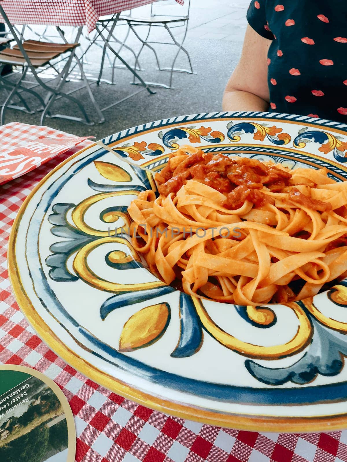 A vibrant plate of fresh pasta topped with rich tomato sauce sits on a checked tablecloth at an outdoor cafe in Italy. Surrounding tables and decor create a charming atmosphere.