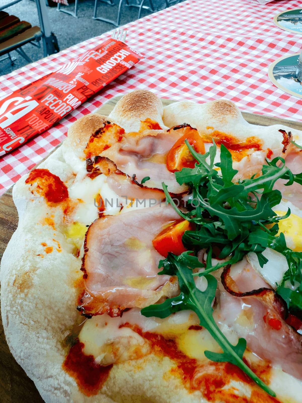 A freshly baked pizza sits on a wooden board, topped with ham, fresh tomatoes, and arugula. The vibrant red and white checkered tablecloth adds charm to the sunny outdoor market atmosphere.