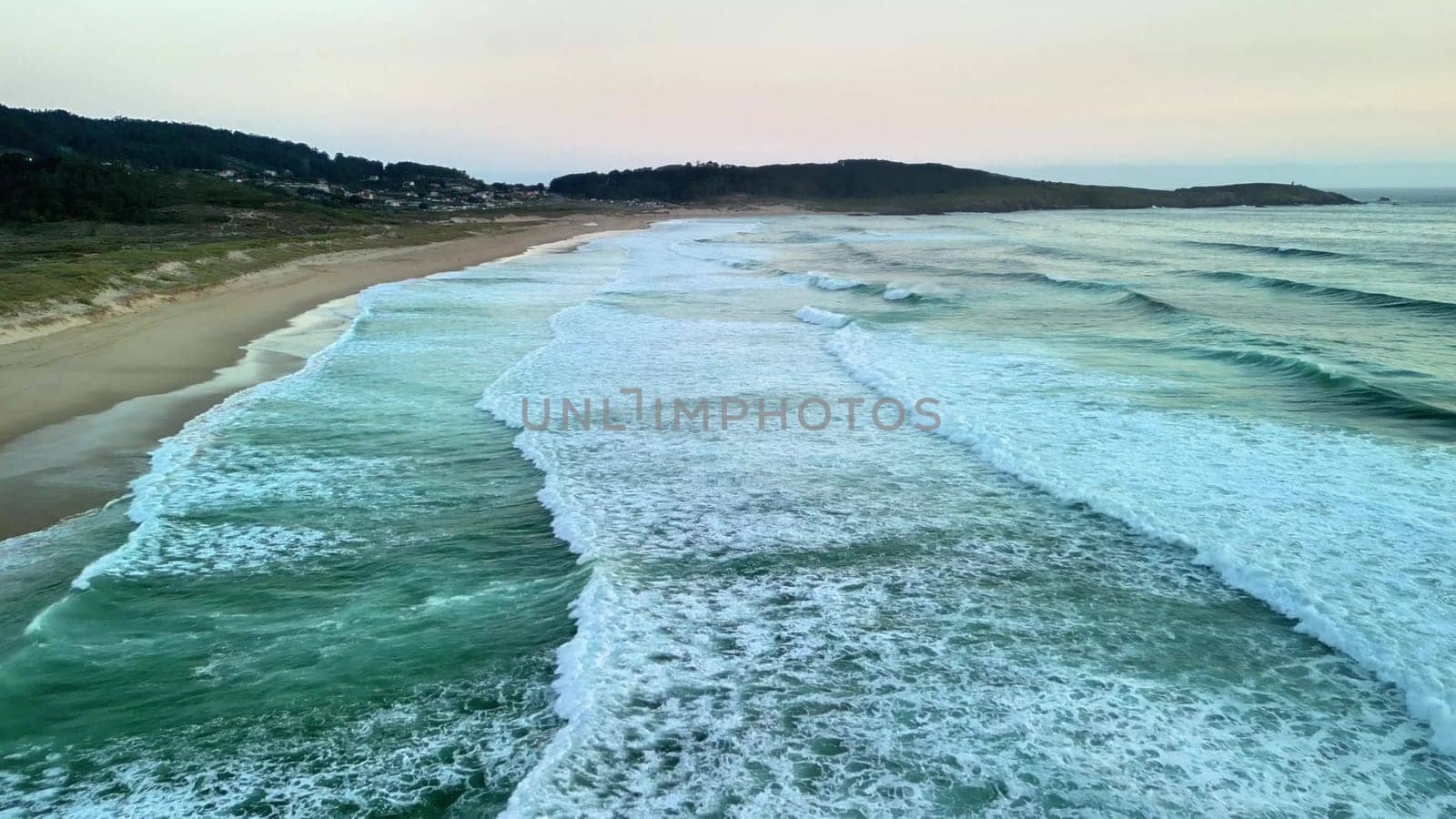 An aerial view shows waves crashing on a tranquil beach at twilight, showcasing the beauty of nature