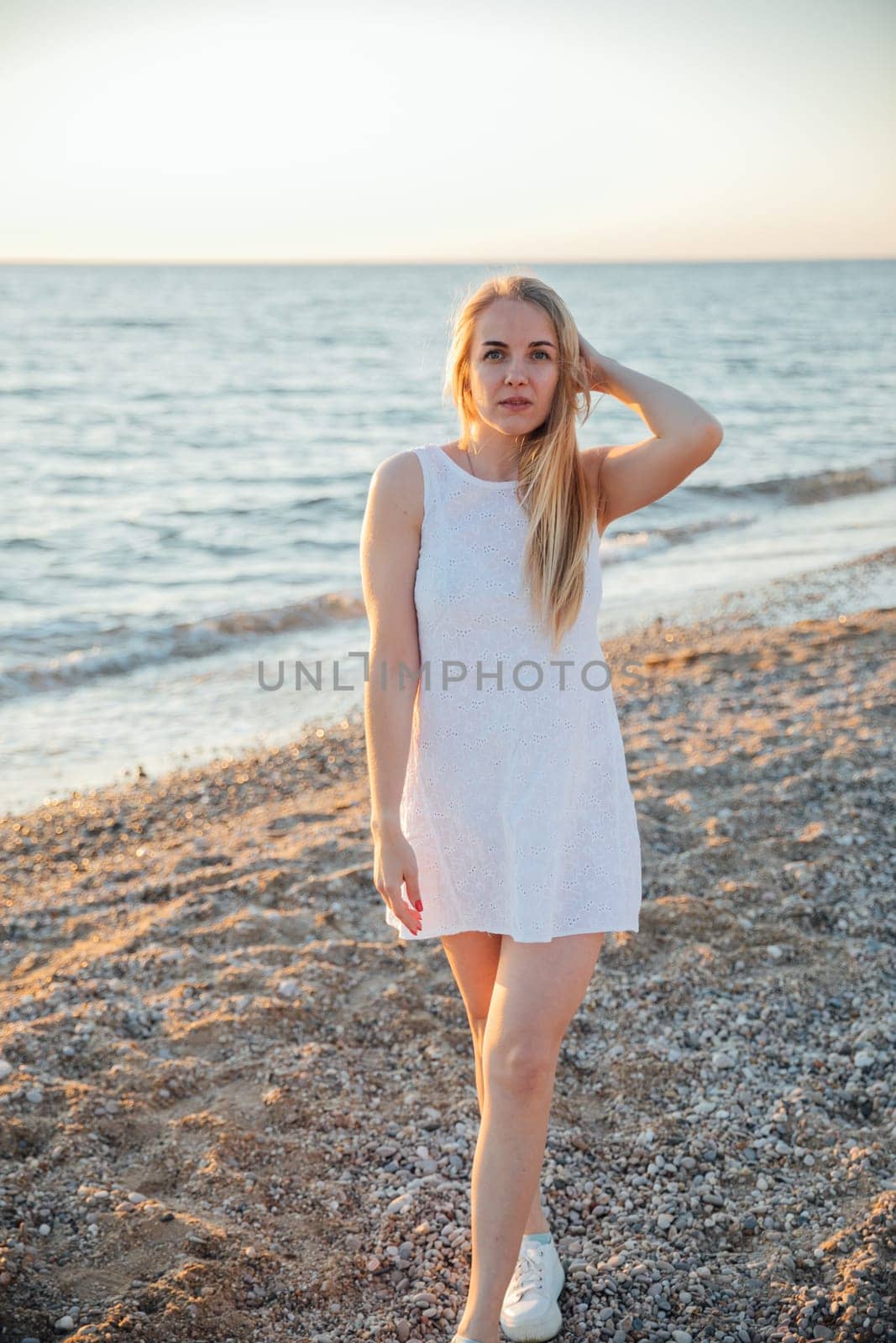 Beautiful blonde woman in summer dress walking on the beach by the sea