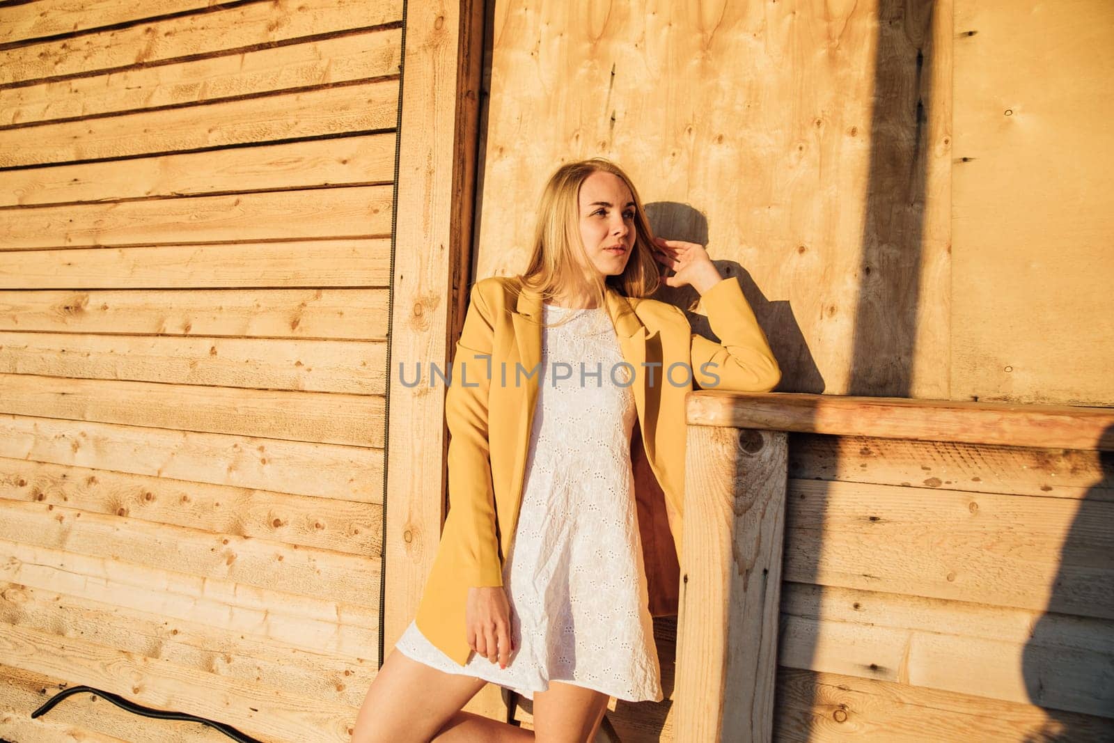 Blonde woman in white dress and yellow jacket sitting near wooden house