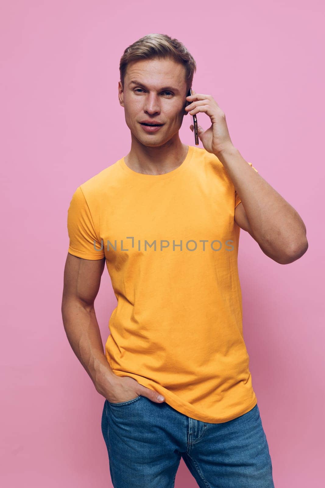 Full body cheerful man engaging in a phone conversation dressed in bright yellow t-shirt and jeans, standing against a pink studio background, expressing friendliness and ease. Concept: communication and positivity
