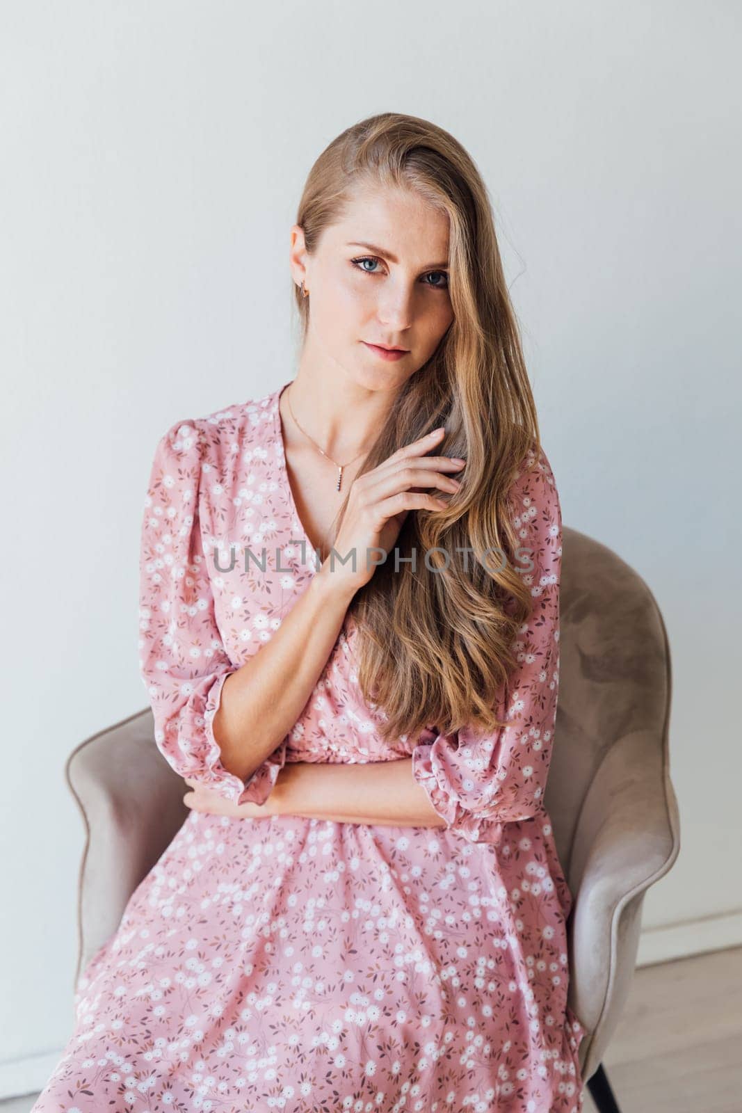 Blonde woman in summer pink dress sitting on chair in white room