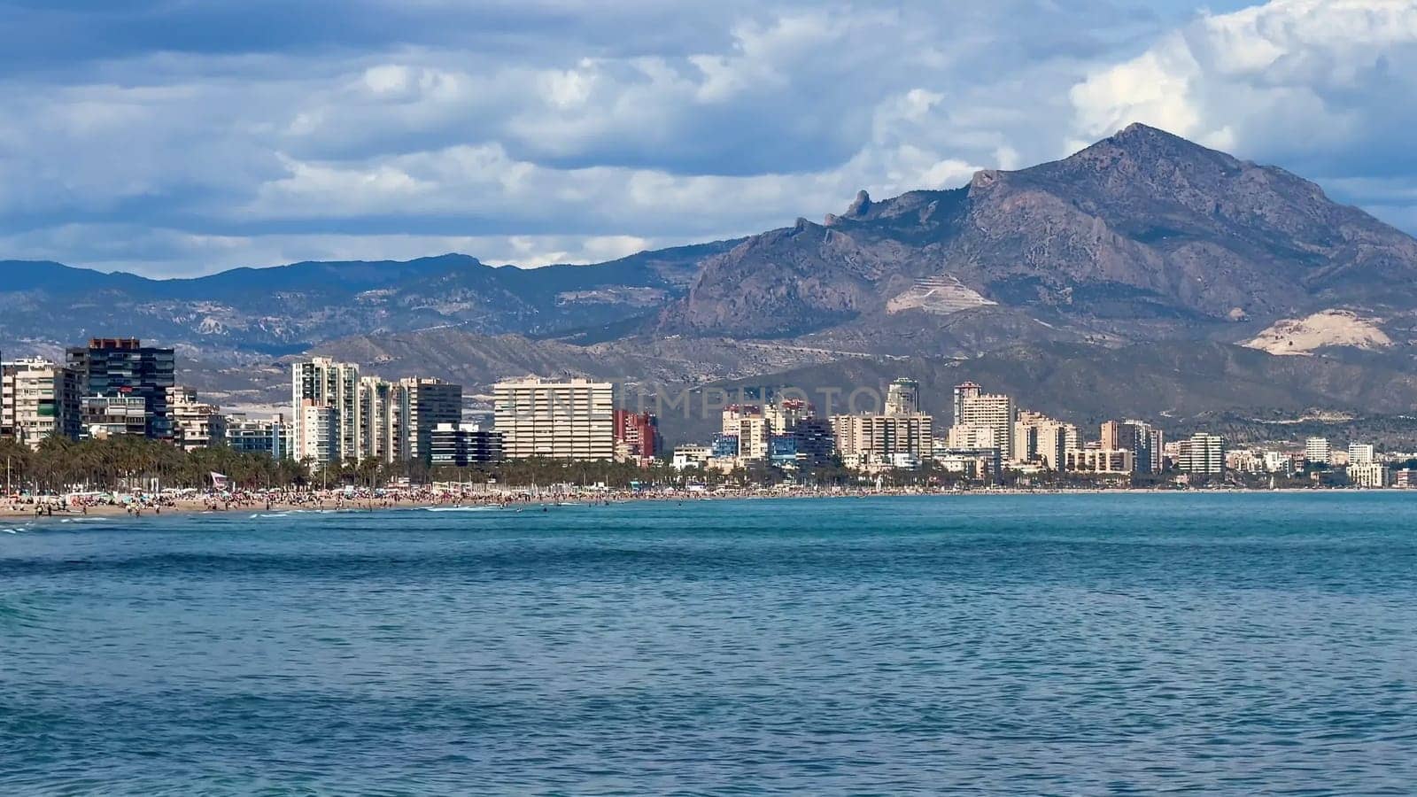 A stunning coastline features modern architecture alongside majestic mountains under a vibrant blue sky