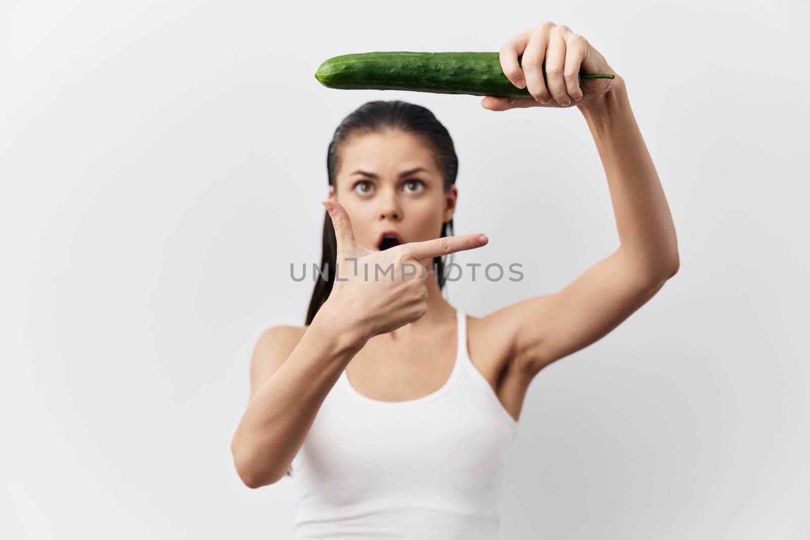 shocked woman pointing finger at cucumber while holding it above her head, displaying an expression of surprise, set against a light neutral background, capturing humor and creativity