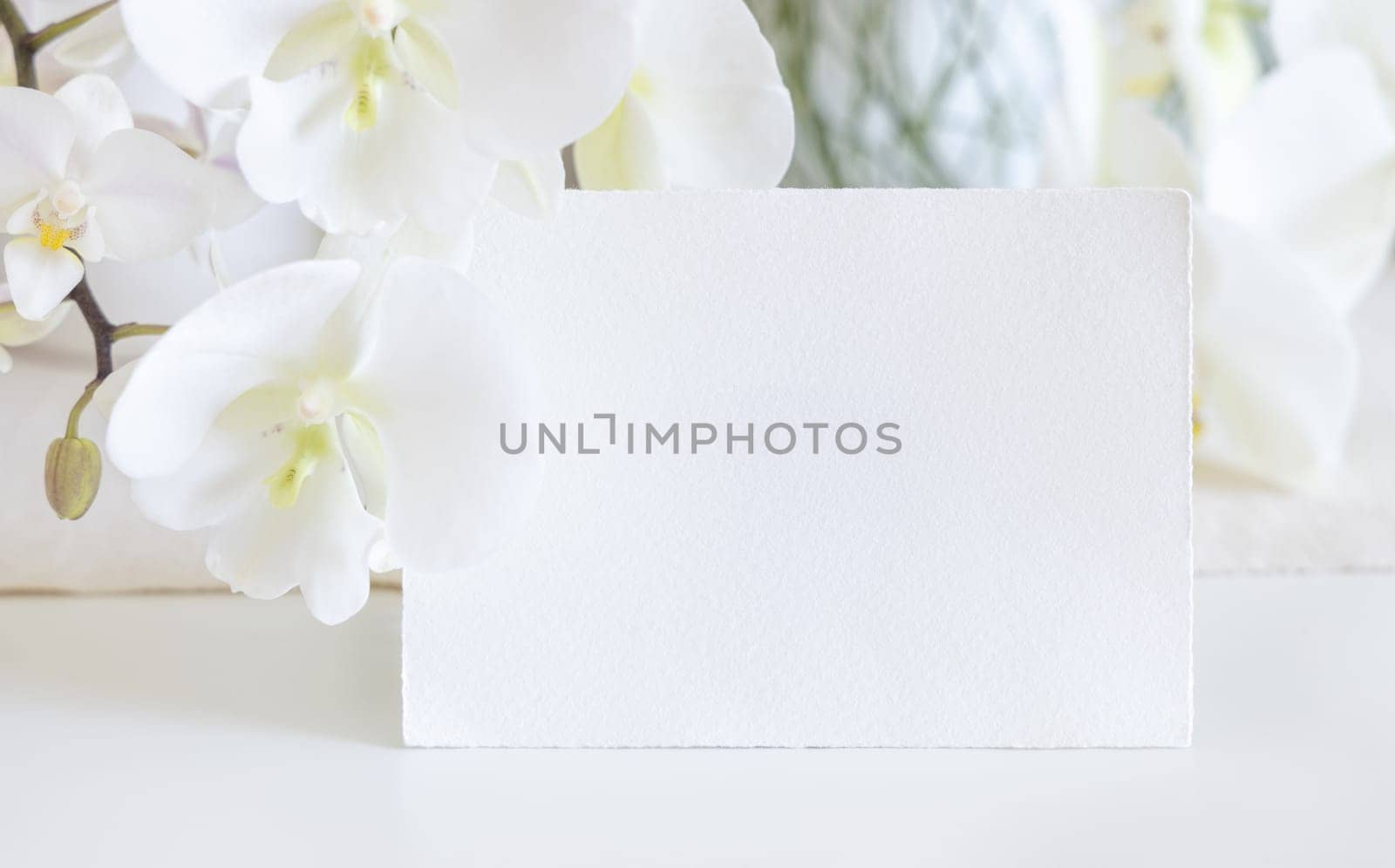 Blank card near white orchid flowers on table close up, mockup. Pastel romantic scene with horizontal card for exotic wedding, Valentines or Mothers day stationery and tropical party design