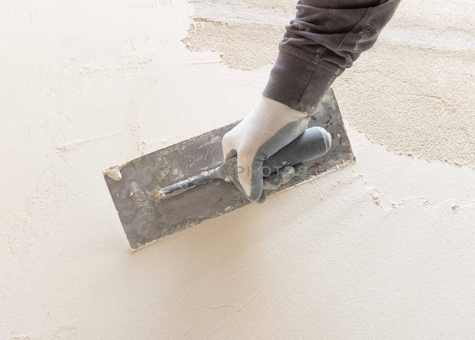 Craftsman uses a float trowel and cement to plaster a house wall. Young men with glasses in gray work clothing during construction or renovation process