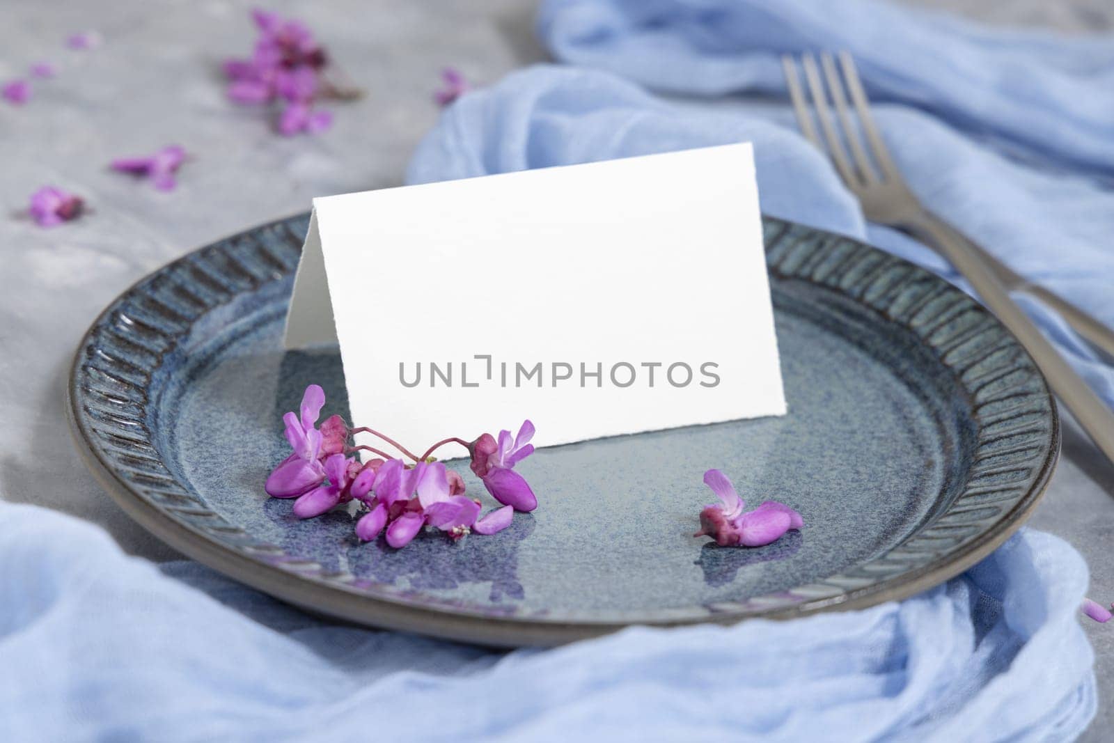 Pastel wedding table place with an folded card on plate near wild purple flowers and light blue fabric closeup. Name card mockup for spring party design or romantic wedding stationery