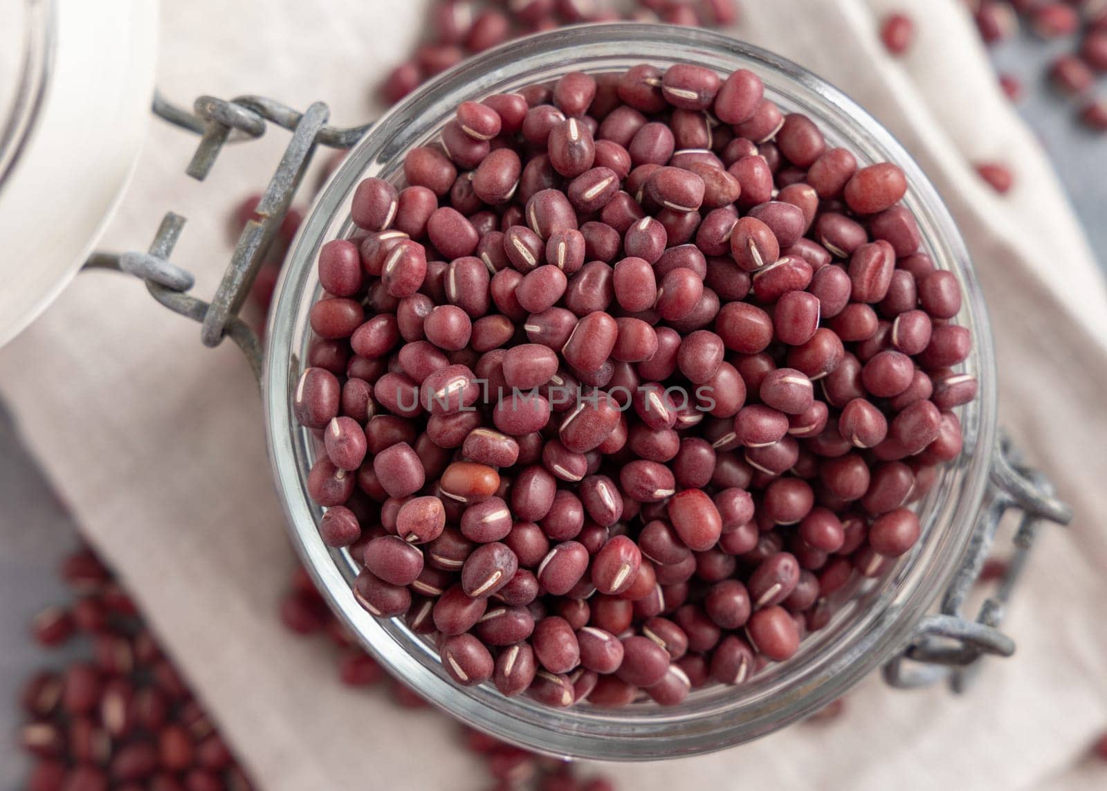 Opened glass jar full of dried red azuki beans on folded napkin on gray table top view. Vegetarian protein source from legumes. Traditional East Asian cousin ingredient