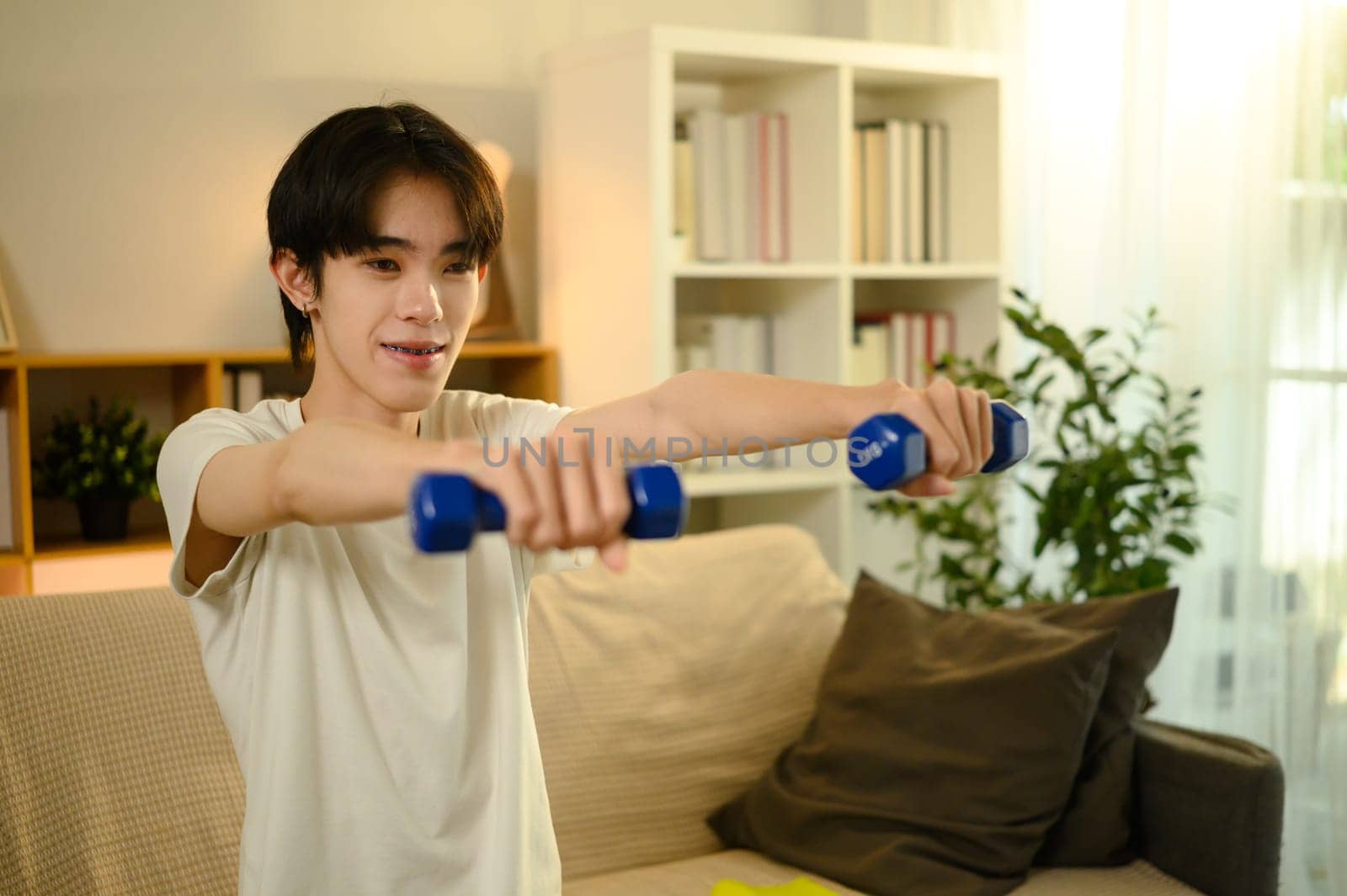 Smiling teenage man lifting blue dumbbells during a home workout.