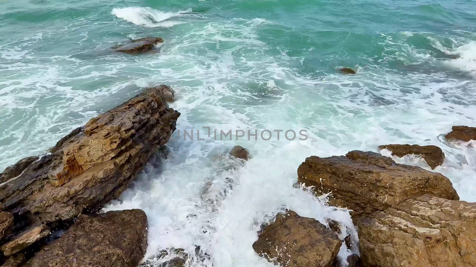 Waves gently crash and collide against the rugged, weatherworn rocks along a tranquil, turquoise coastline
