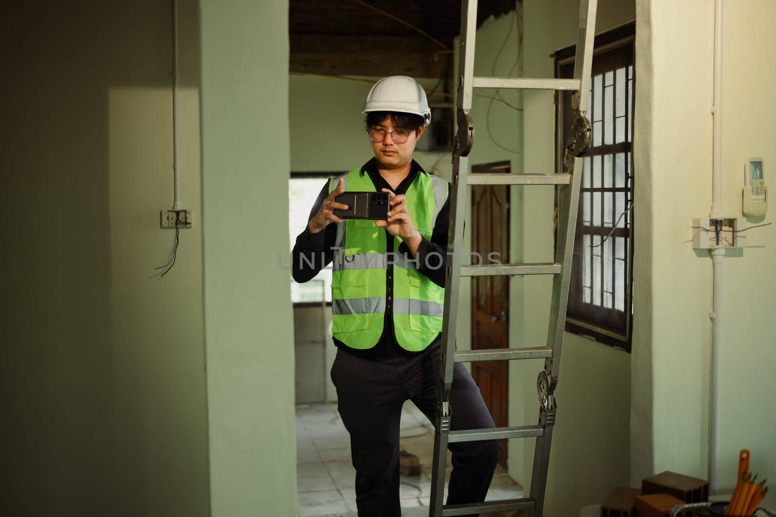Engineer in safety vest and hard hat photographing project progress with smartphone.
