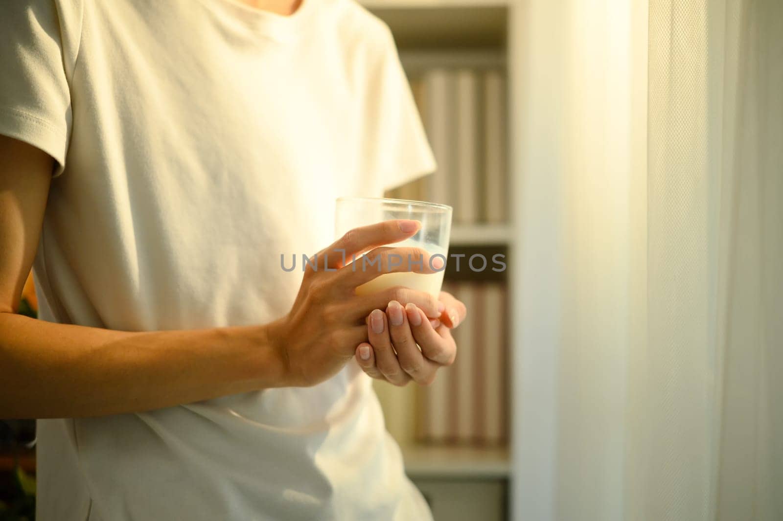 Close up of a man holding a glass of milk while standing indoors in warm natural light.