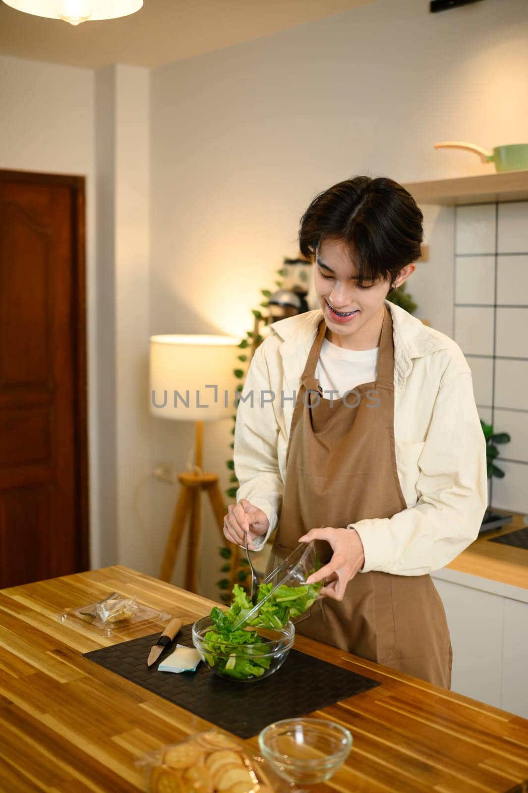 Smiling male in apron preparing a fresh green salad in a modern kitchen.