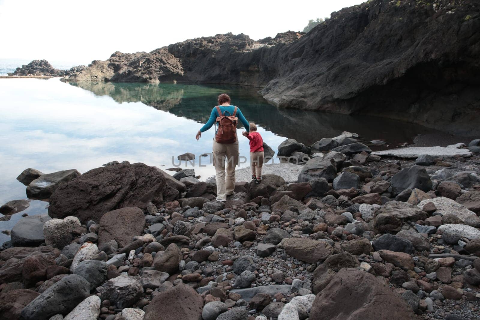 Mother and child carefully walking on rocky terrain near calm water, enjoying a hiking adventure along a scenic coastline