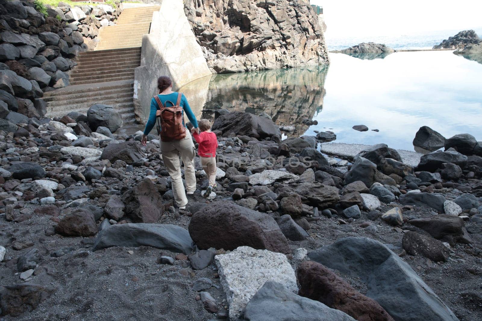 Mother and child exploring rocky beach near volcanic cliffs and calm water