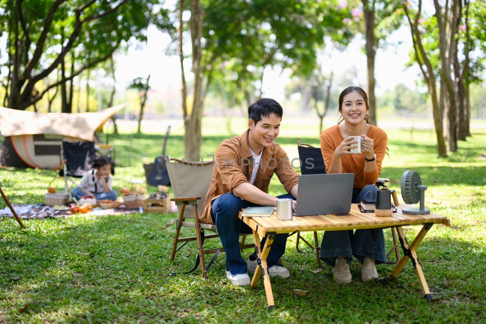 Happy couple using laptop while camping with their daughter in the background.