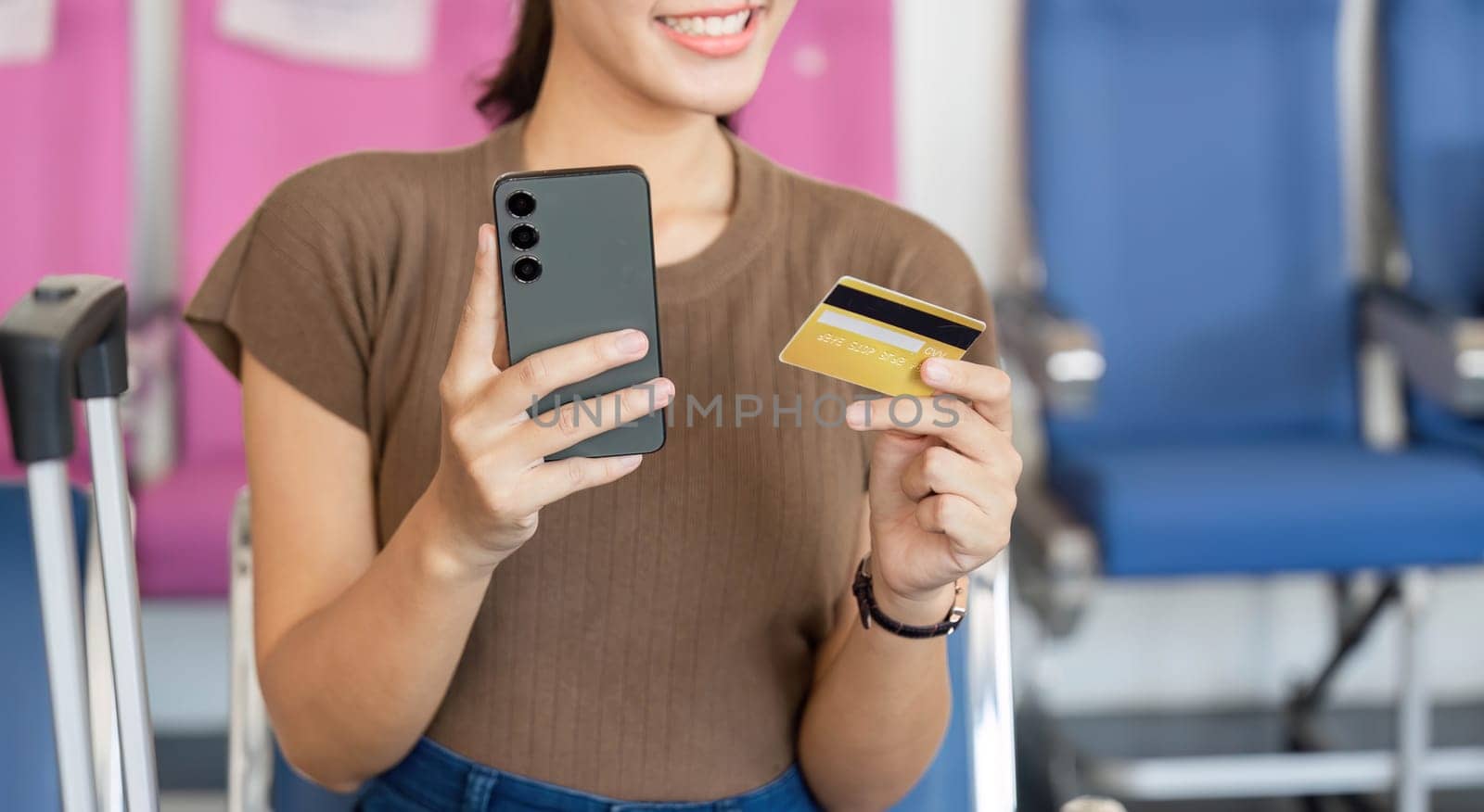 A young woman smiling while holding a smartphone and a credit card, preparing for her travel at the airport, highlighting excitement and anticipation.