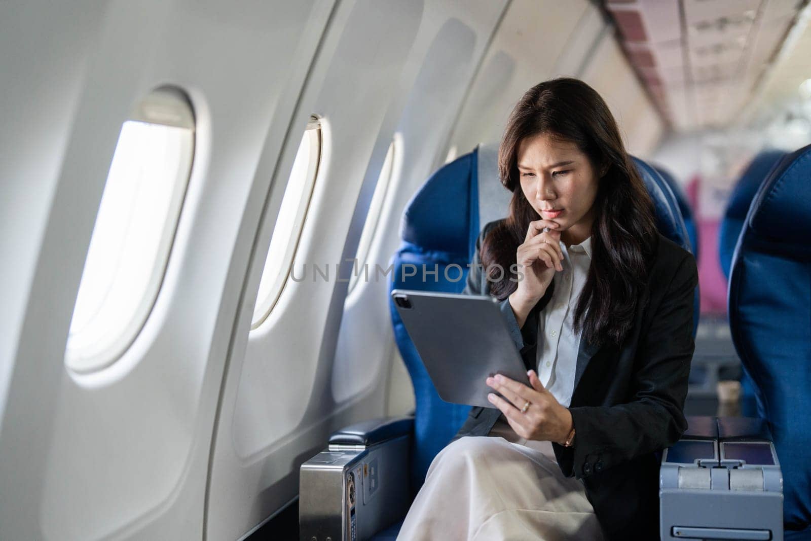 A young businesswoman looking concerned while using a tablet on an airplane, deep in thought about her plans.