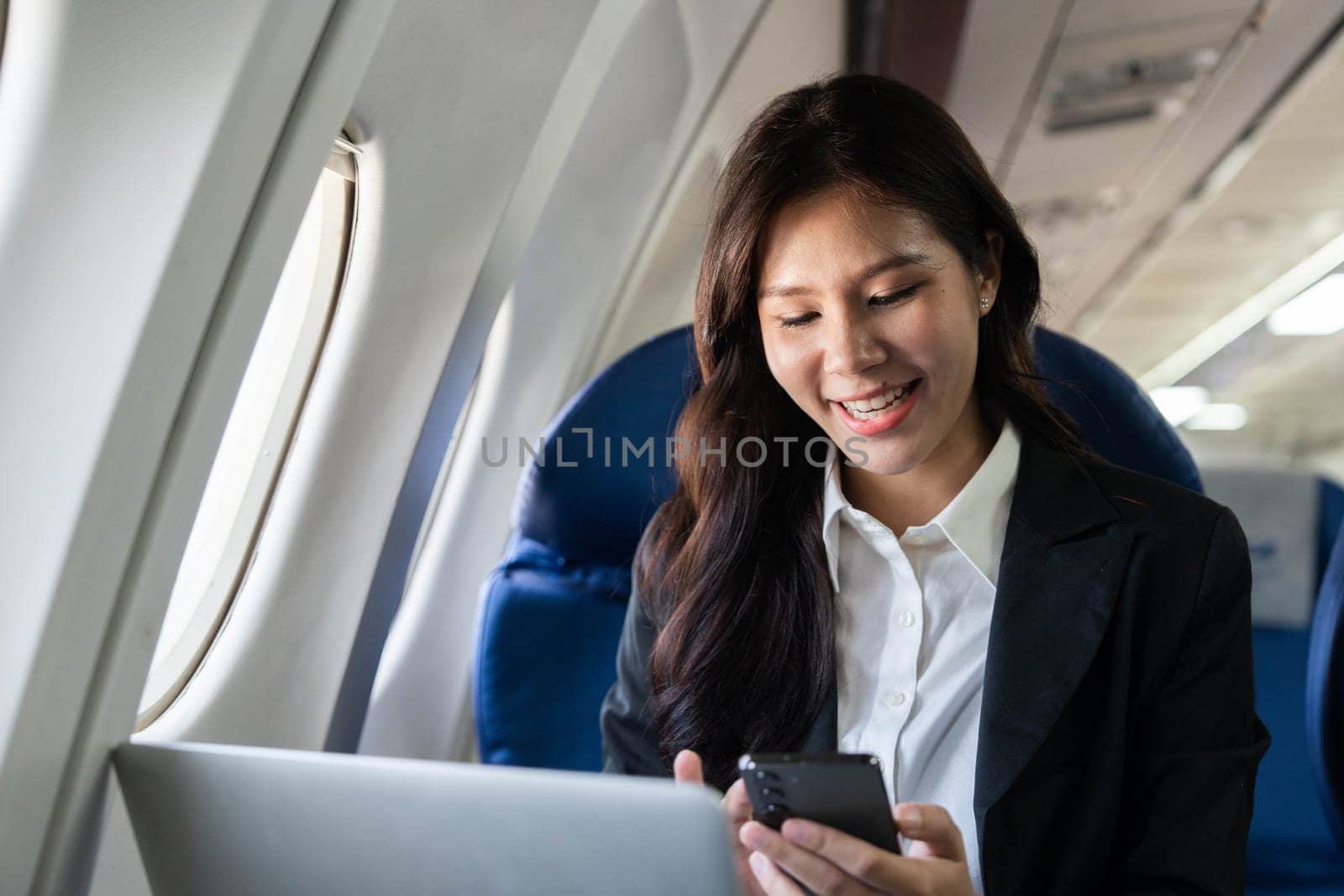 On her flight, a smiling businesswoman enjoys using her phone and laptop, reflecting a balanced approach to work and travel.
