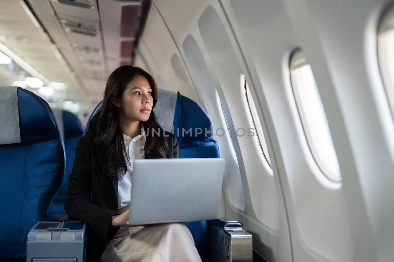 A professional woman reflecting while working on a laptop, looking out of the airplane window during her business travel.