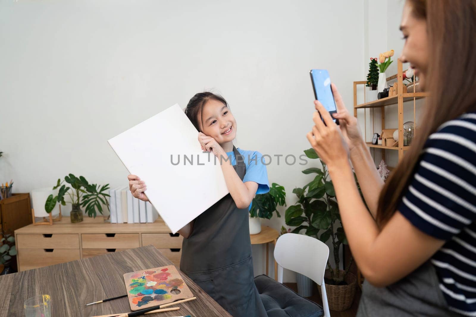 A mother captures a joyful moment of her daughter holding a canvas during family time, celebrating creativity in an art studio.