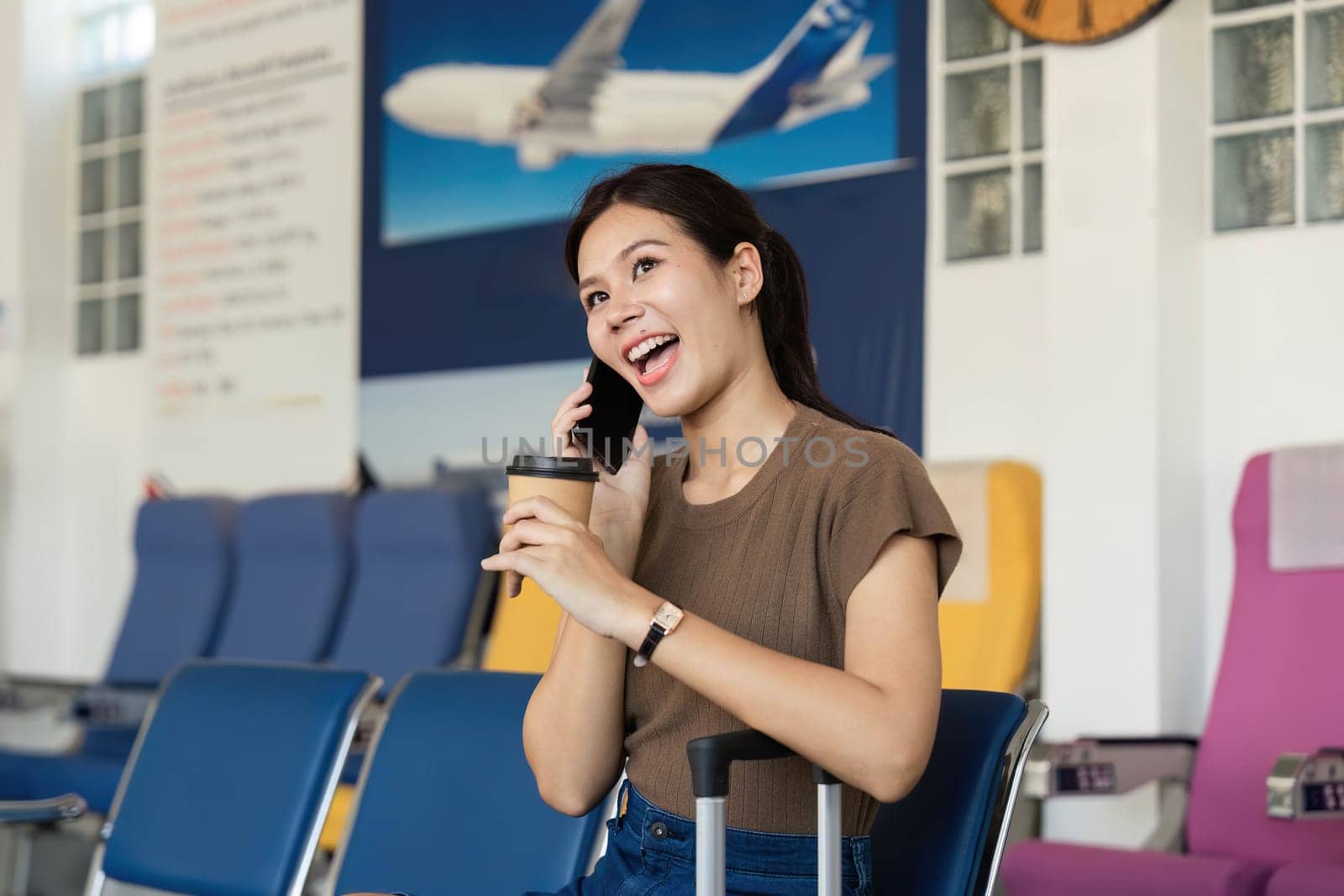 A cheerful young woman at the airport sipping coffee while chatting on her phone, ready for her travel adventure.