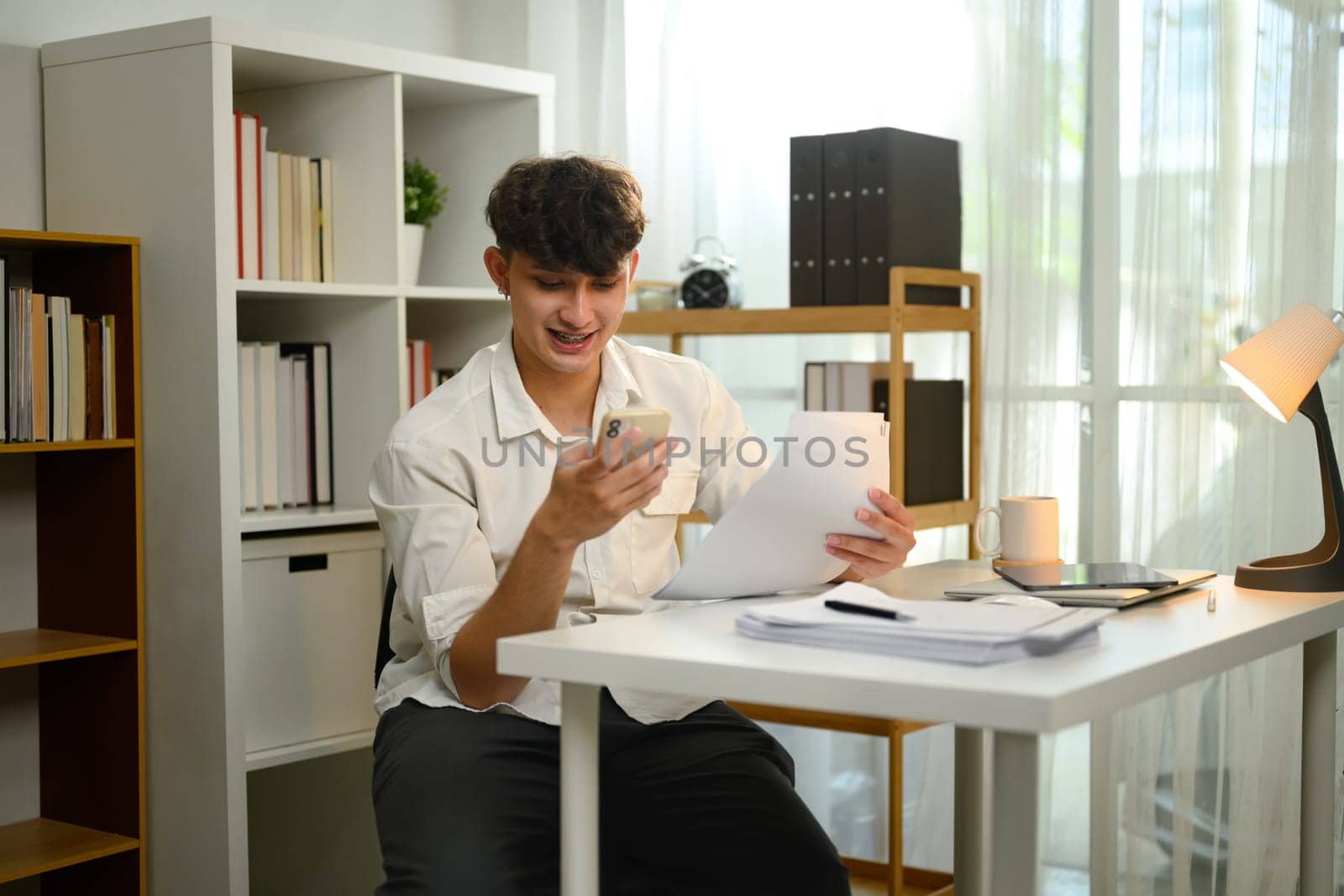 Smiling man using smartphone while reviewing documents in a home office.