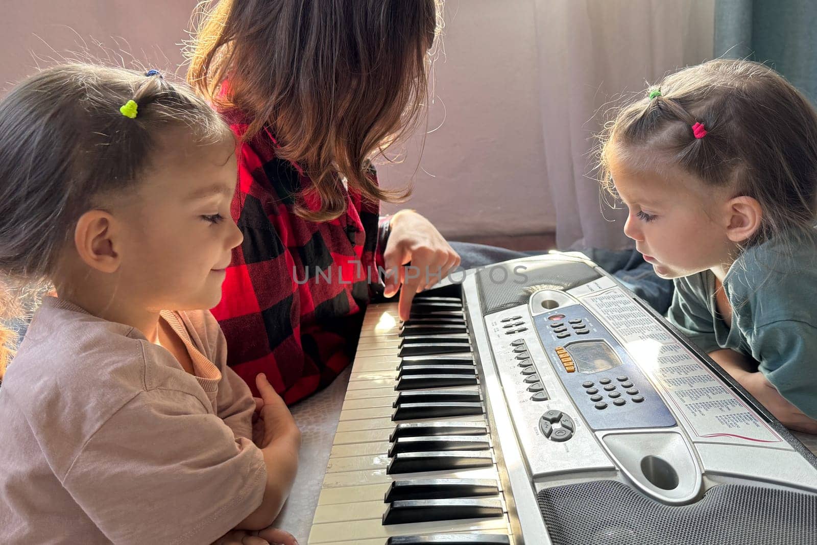 Teen boy shows his twin toddler sisters how to use a synthesizer. The kids sit on the floor, laughing and learning through music. Happy family bonding indoors real life.