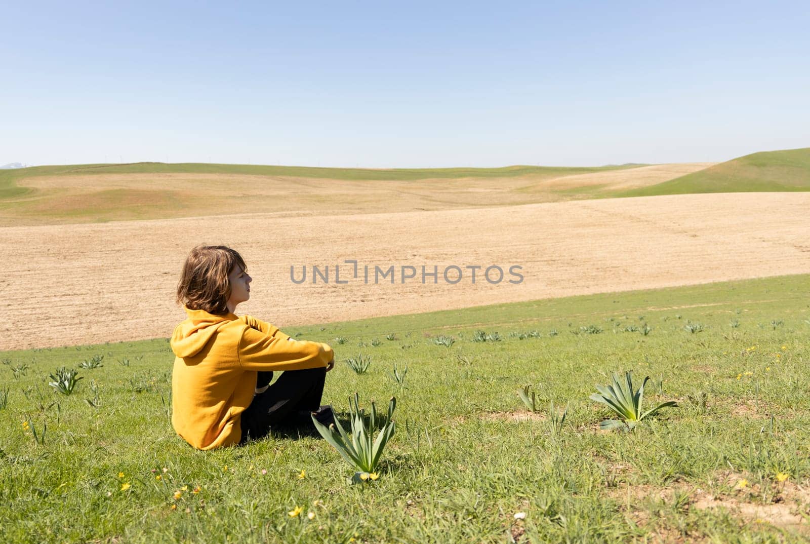 Side view of a person sitting on green grass in open field, looking into the distance. Peaceful outdoor scene with copy space for text or design.