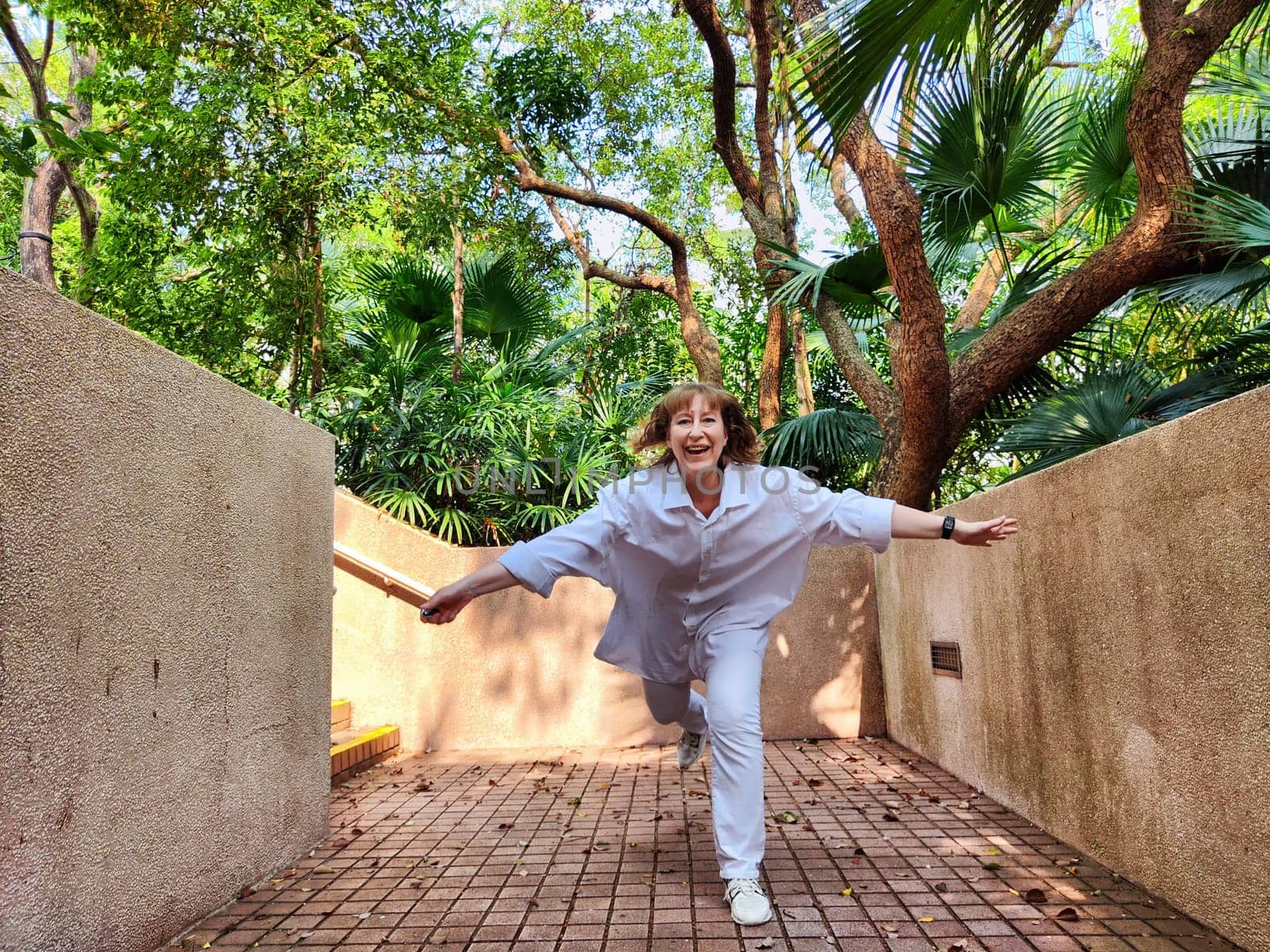A mature blonde woman joyfully poses with arms outstretched in a vibrant tropical garden, embracing the thrill of her travel adventure.