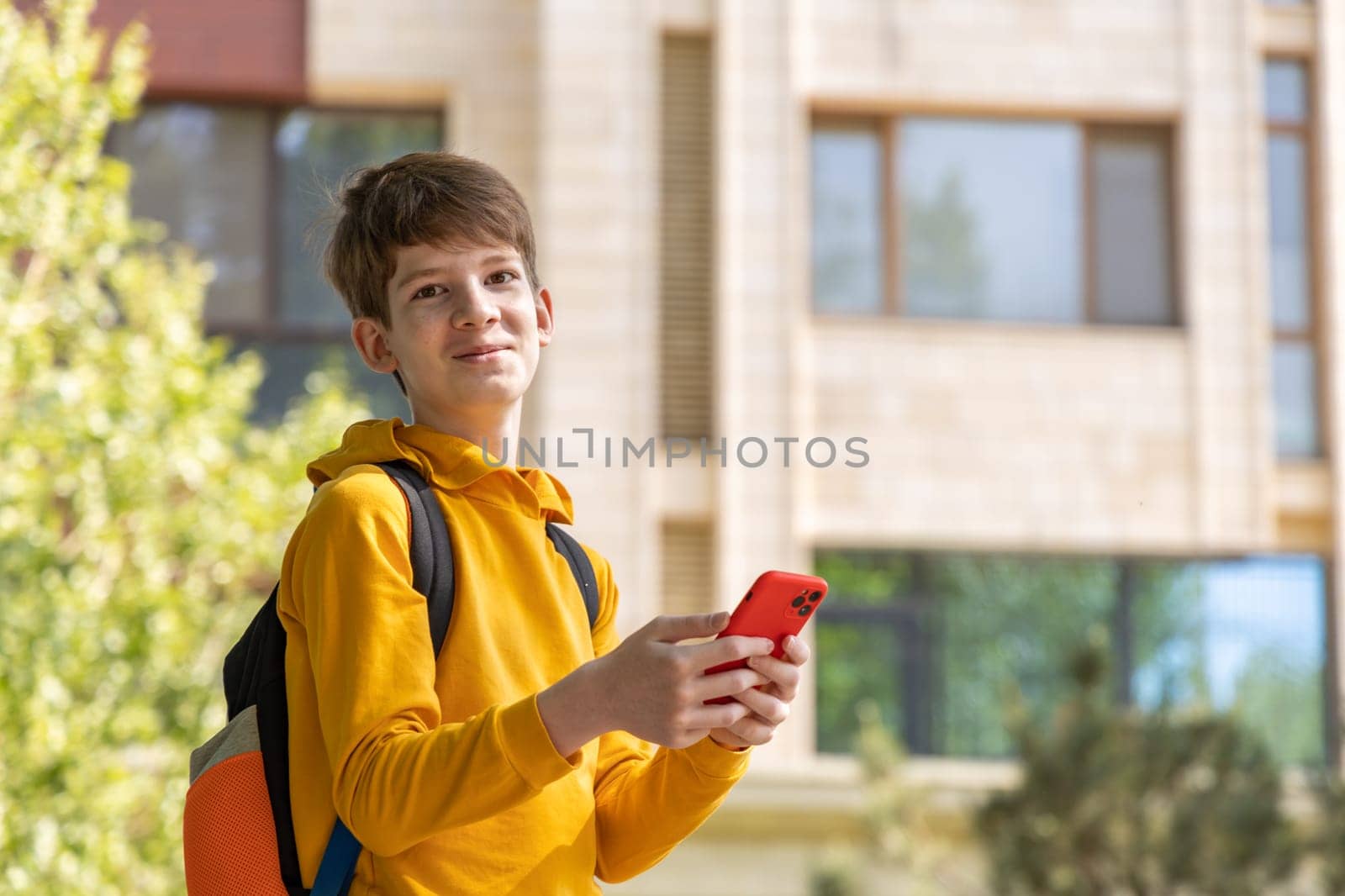 Smiling teenage boy holding smartphone, standing outdoors in front of modern new house. Youth, technology, and contemporary suburban lifestyle concept.