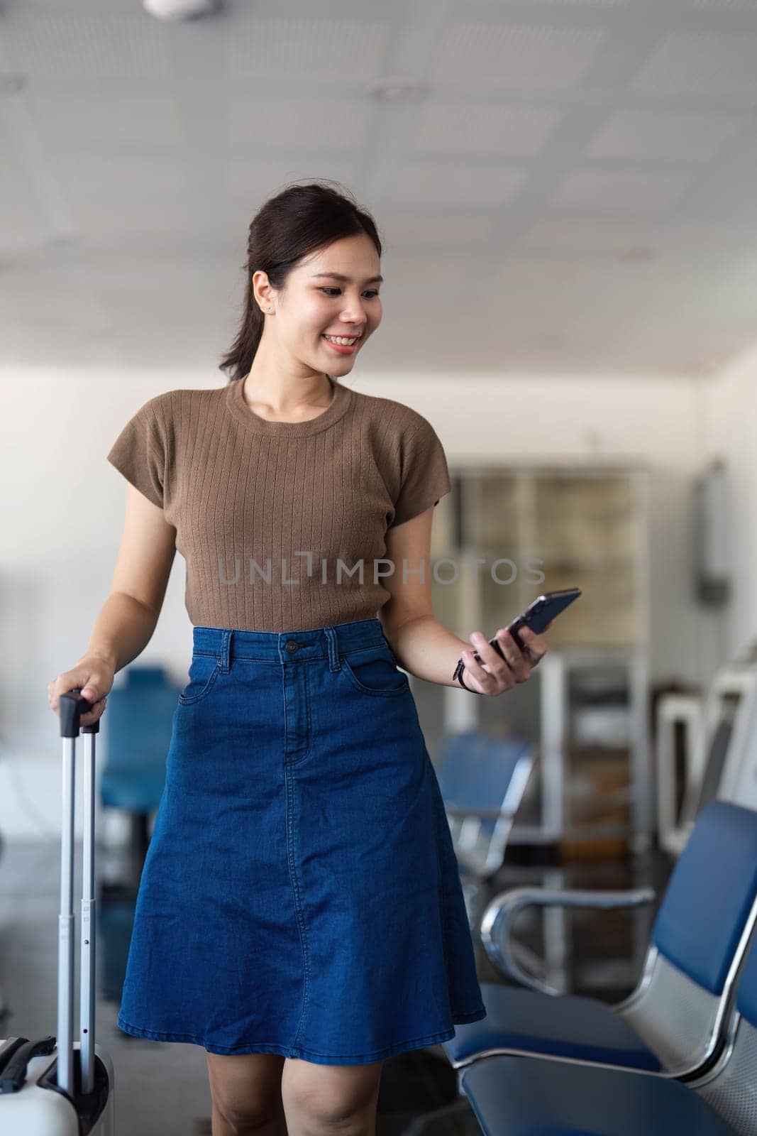 A young woman at the airport smiling while pulling her suitcase and looking at her phone, ready for her next adventure.