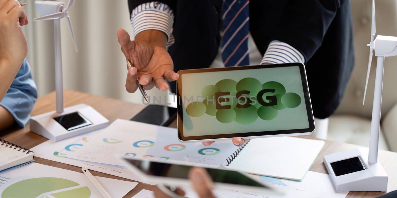 A professional presenting ESG data results on a tablet while discussing renewable energy technologies using wind turbine models during a meeting.